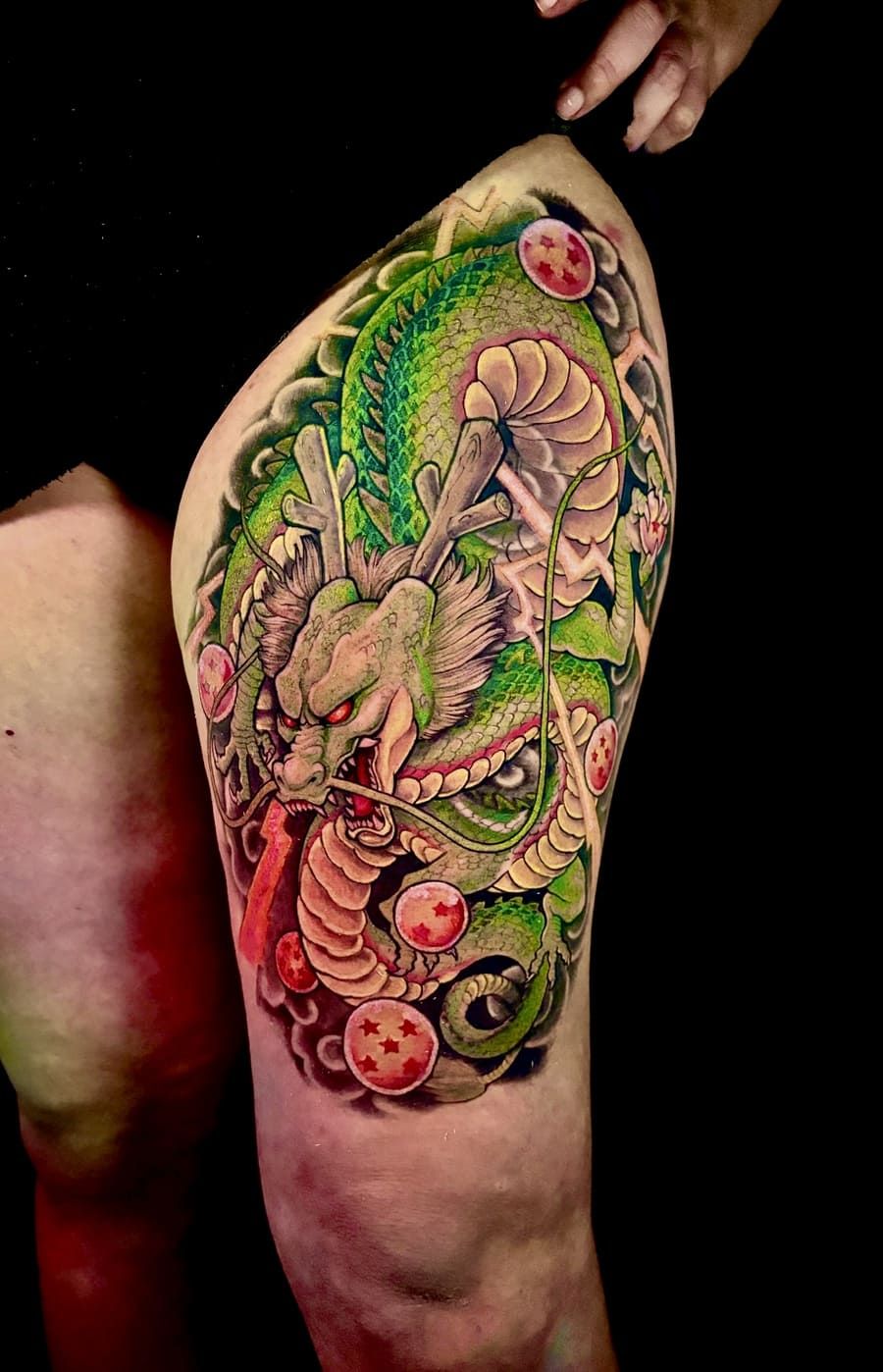 Tatuaje de Dragon Ball Z en un muslo; dragón verde, ojos rojos, sosteniendo bolas de dragón, detallado.