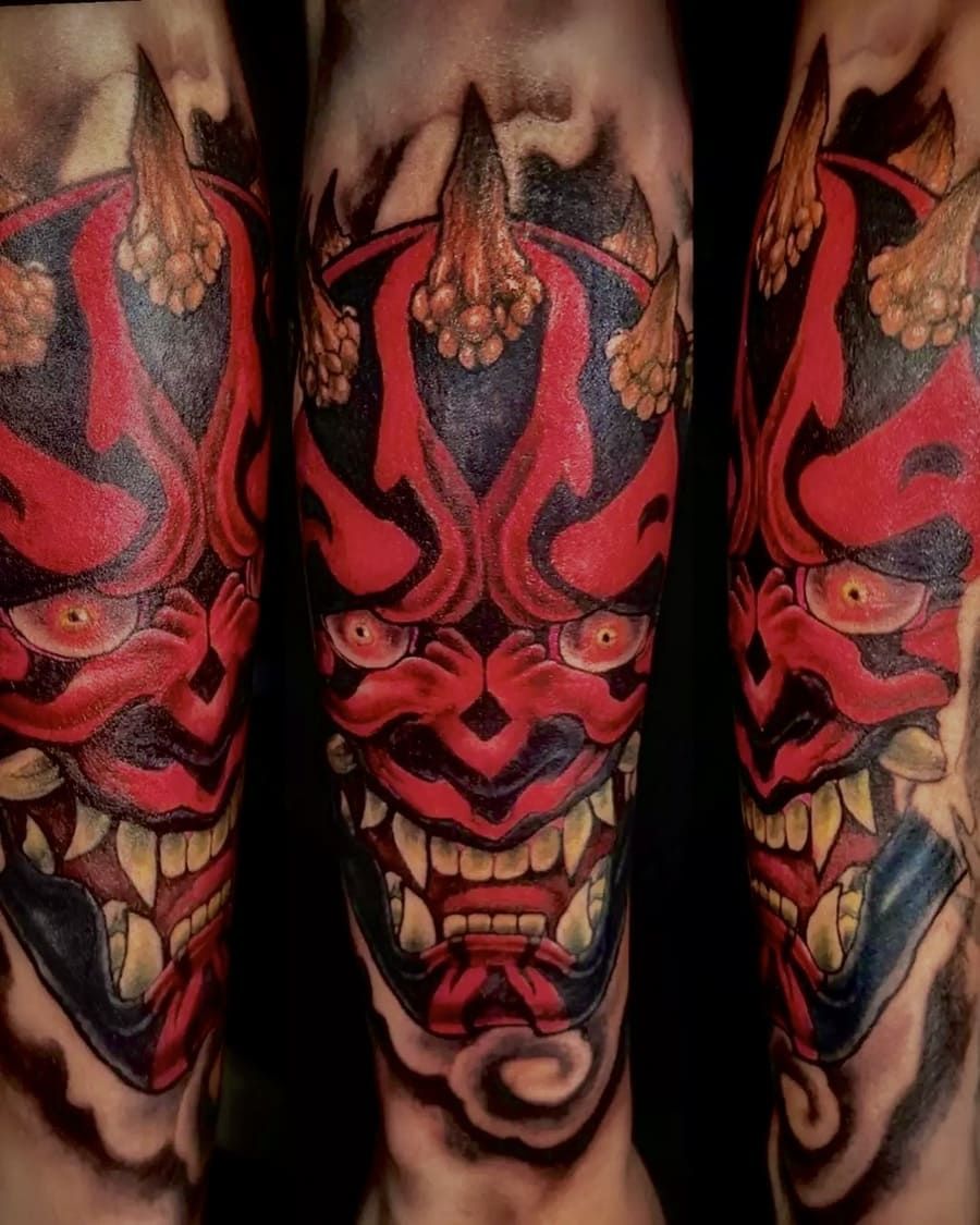 Tatuaje de una máscara Oni roja y negra con dientes afilados, cuernos y ojos amenazantes, en un brazo.