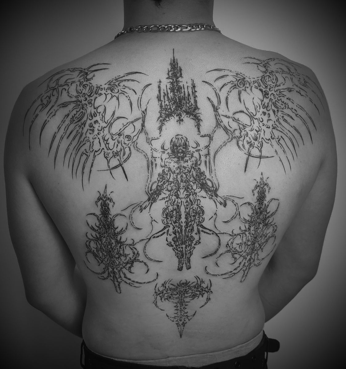 Tatuaje en la espalda en blanco y negro: diseño intrincado de estilo gótico con una figura central, alas y elementos arquitectónicos.