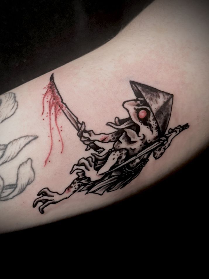 Tatuaje negro y rojo de una rana demoníaca que sostiene un cuchillo ensangrentado y lleva un sombrero cónico.