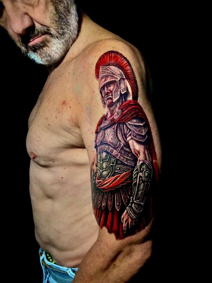 Hombre con un tatuaje detallado de un soldado romano en la parte superior del brazo, vestido con capa y casco rojos.