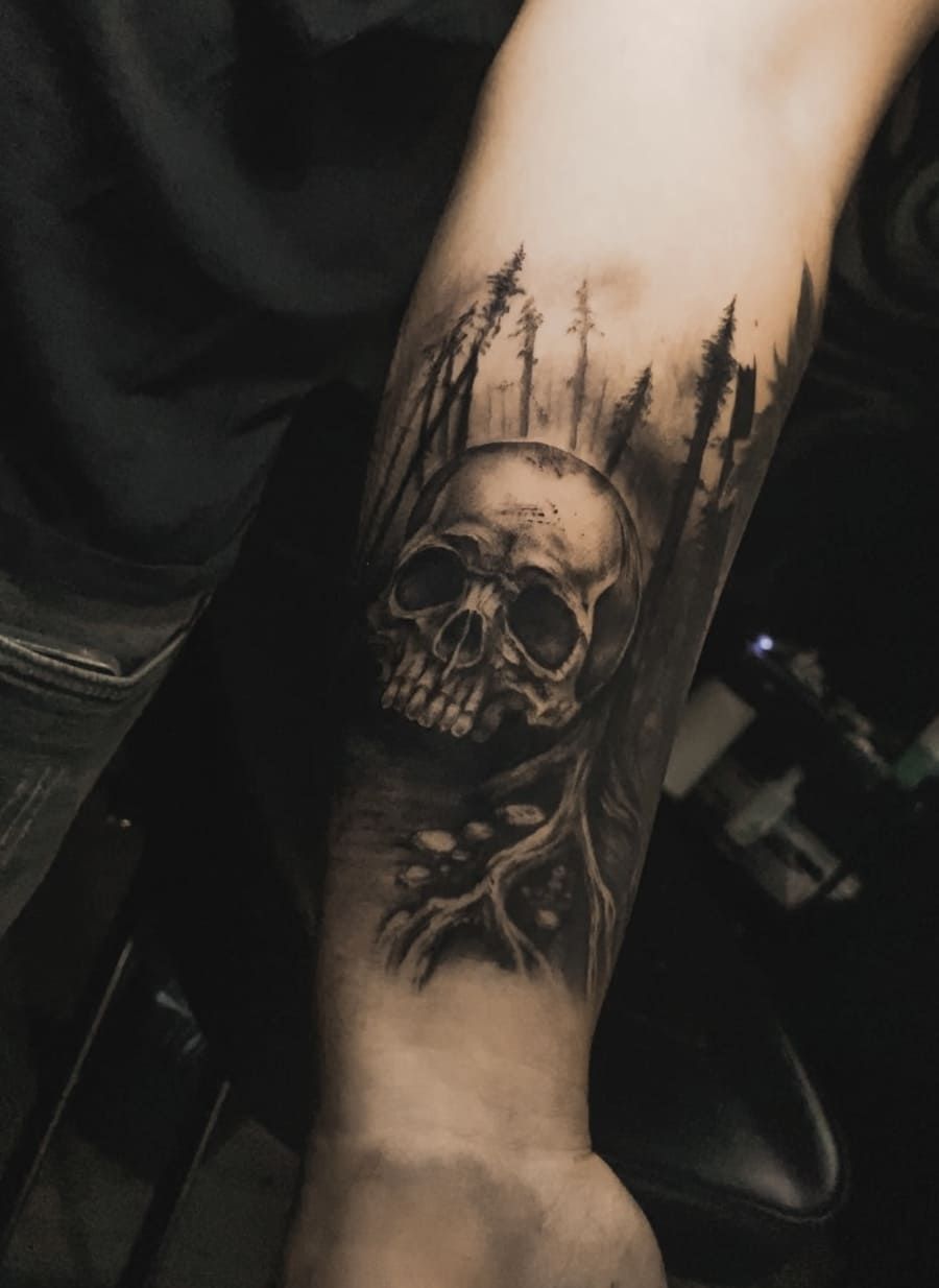 Tatuaje de calavera en un brazo, rodeada de un bosque de árboles en escala de grises.