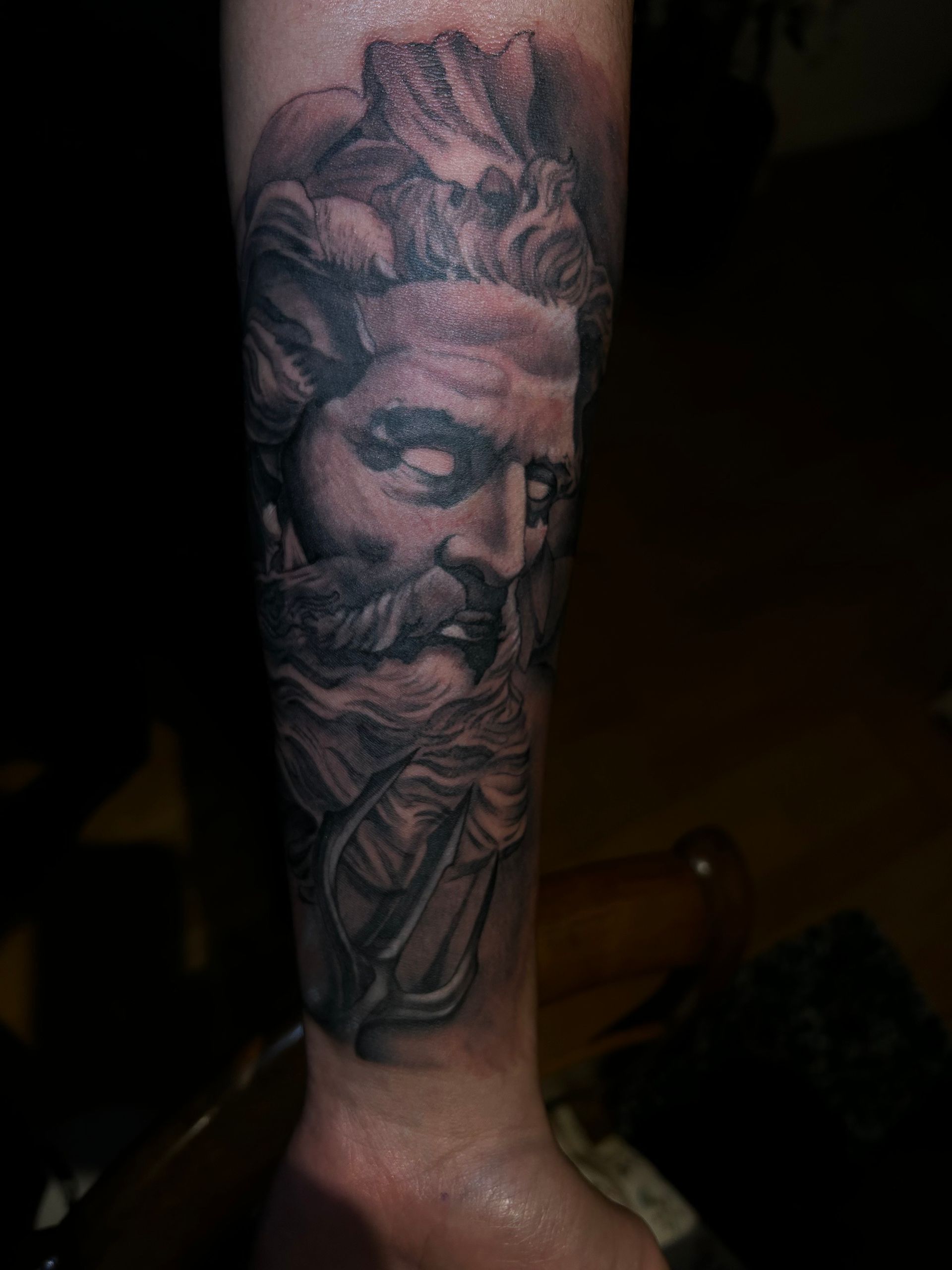 Tatuaje negro y gris de un rostro barbudo, posiblemente Poseidón, sosteniendo un tridente en el antebrazo.