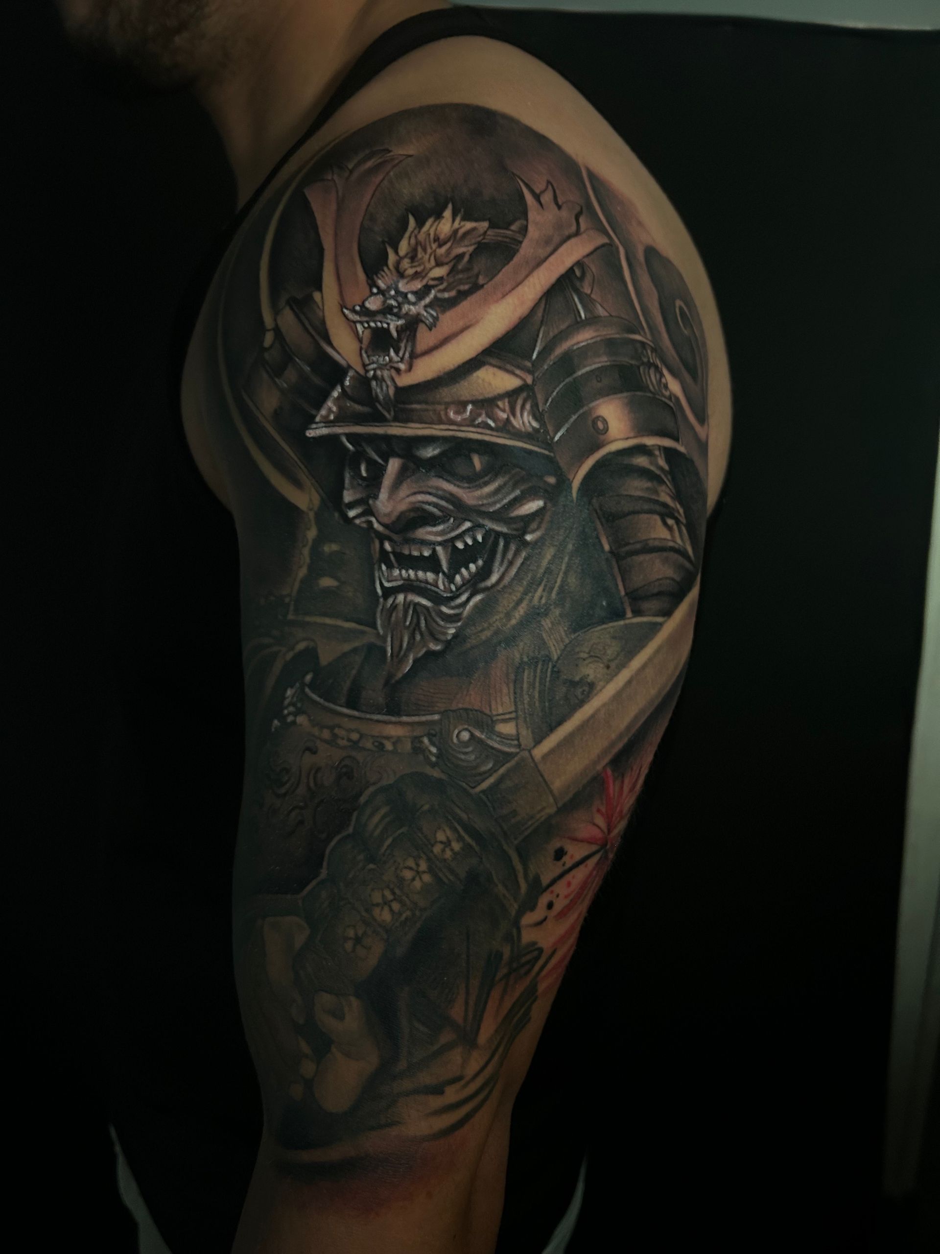 Tatuaje negro y gris de un guerrero samurái en un brazo, con armadura y casco con detalles de dragón.