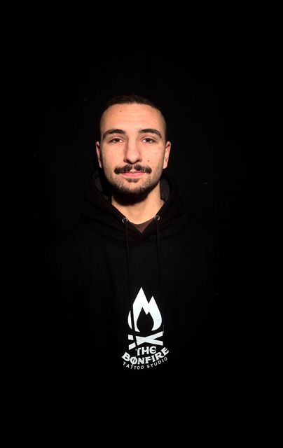 Hombre con los brazos cruzados, vestida con una sudadera negra con el logo de una llama blanca y fondo oscuro.