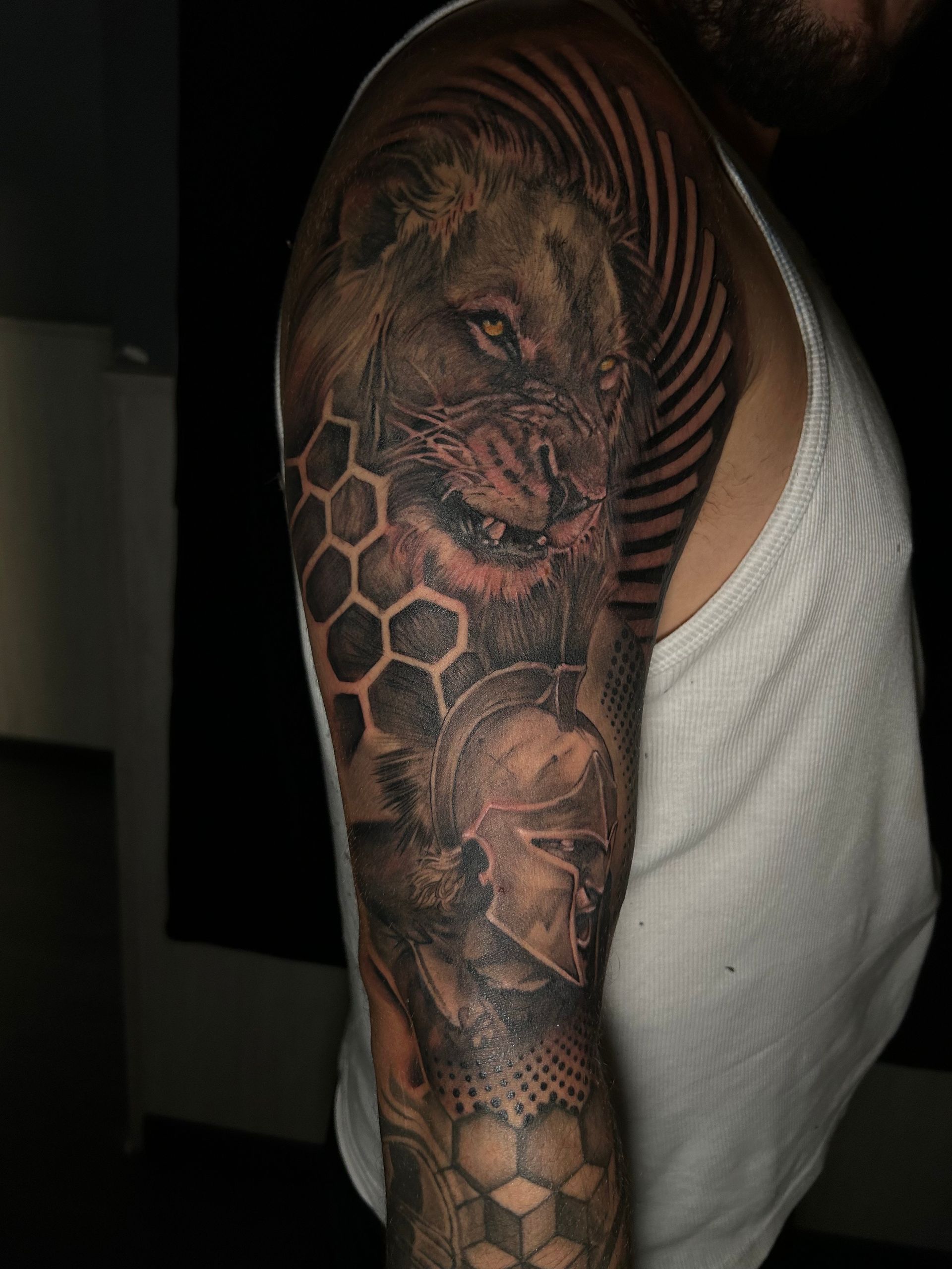 Tatuaje en un brazo que representa un león, un casco y formas de panal; tinta negra y gris.
