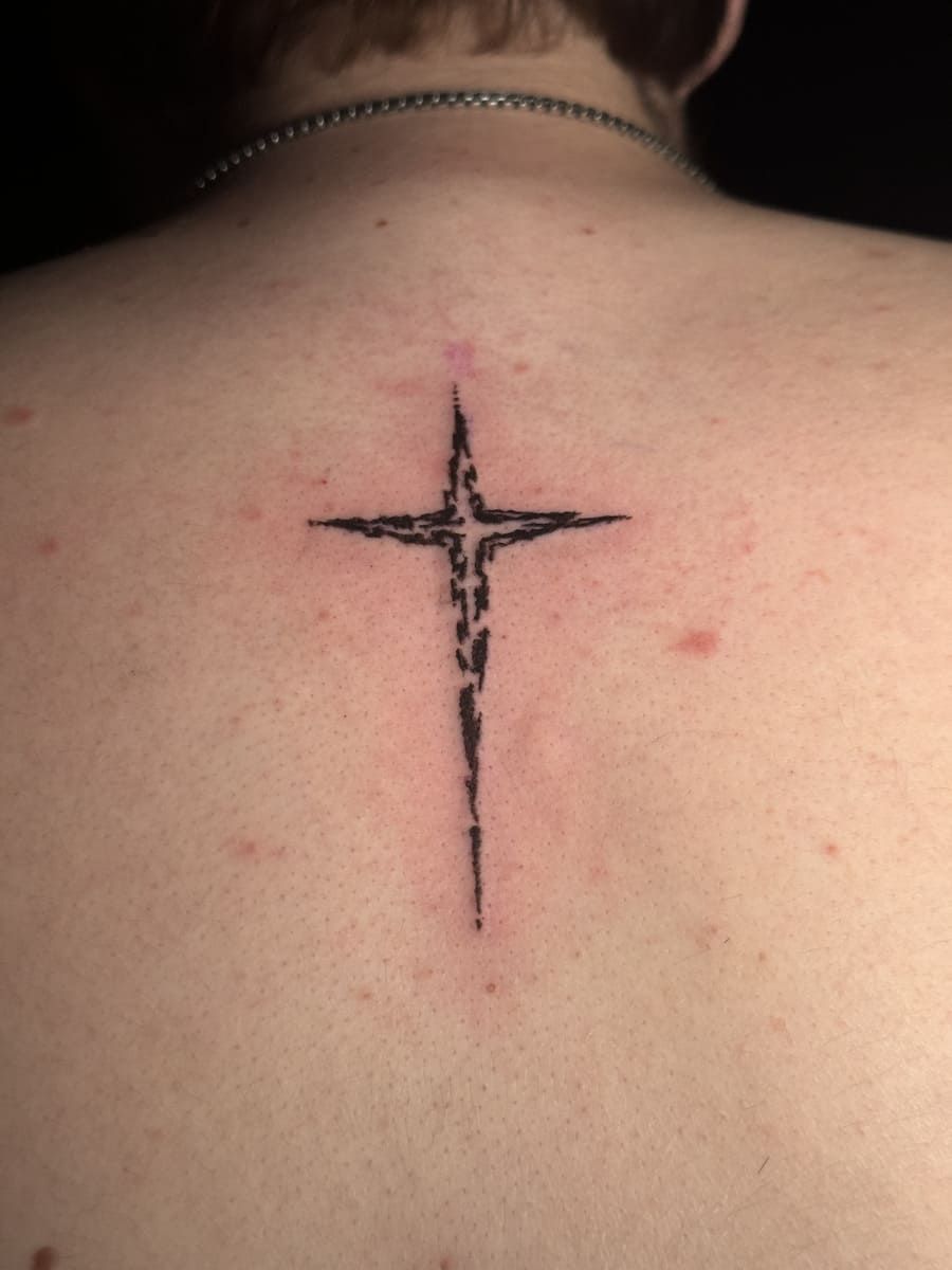 Tatuaje en forma de cruz en la espalda de una persona, tinta negra, bordes irregulares, pequeño collar de cadena visible.