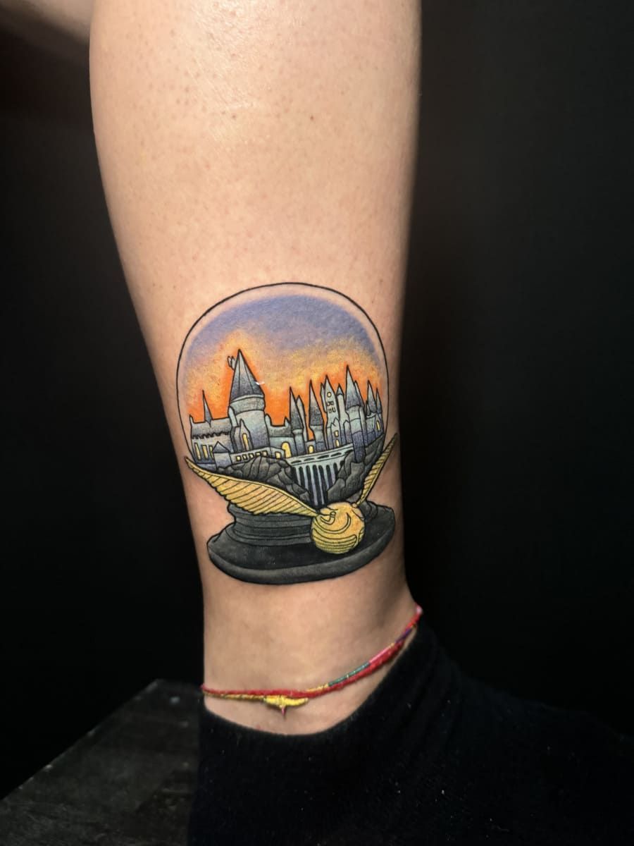 Tatuaje del castillo de Hogwarts dentro de una bola de nieve, con una snitch dorada en la base. Fondo de cielo colorido.