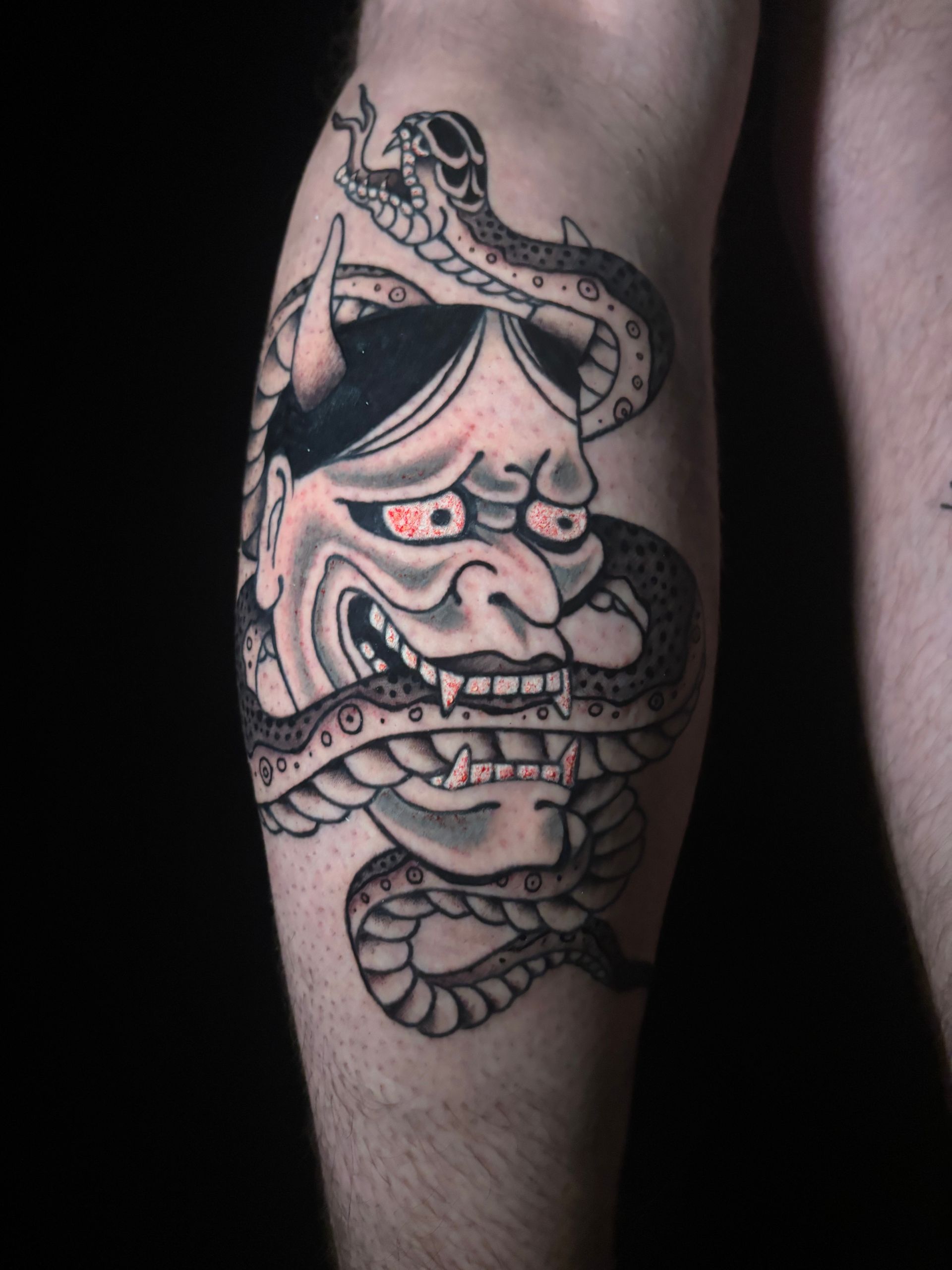 Tatuaje de una máscara de demonio japonesa con ojos rojos, colmillos y una serpiente envuelta a su alrededor en una pierna.