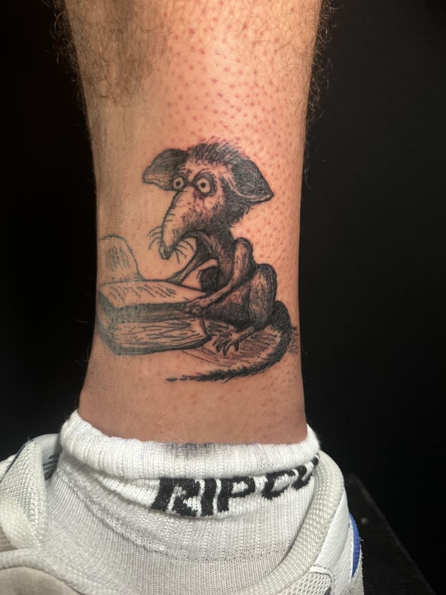 Tatuaje de una rata leyendo un libro en un tobillo, en escala de grises.