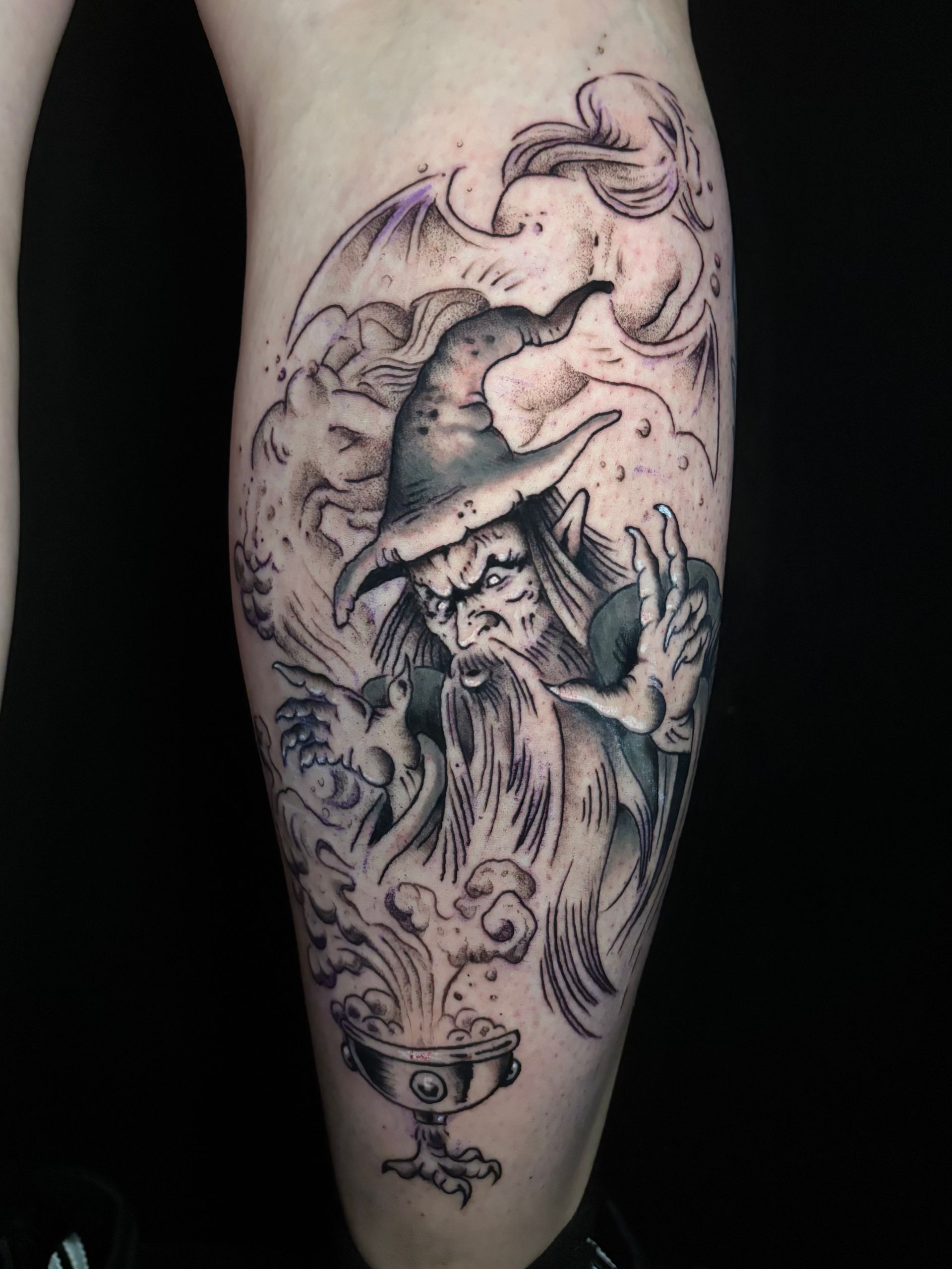 Tatuaje de un mago lanzando un hechizo. El mago lleva sombrero, barba y sostiene un caldero con las manos en una escena nublada.