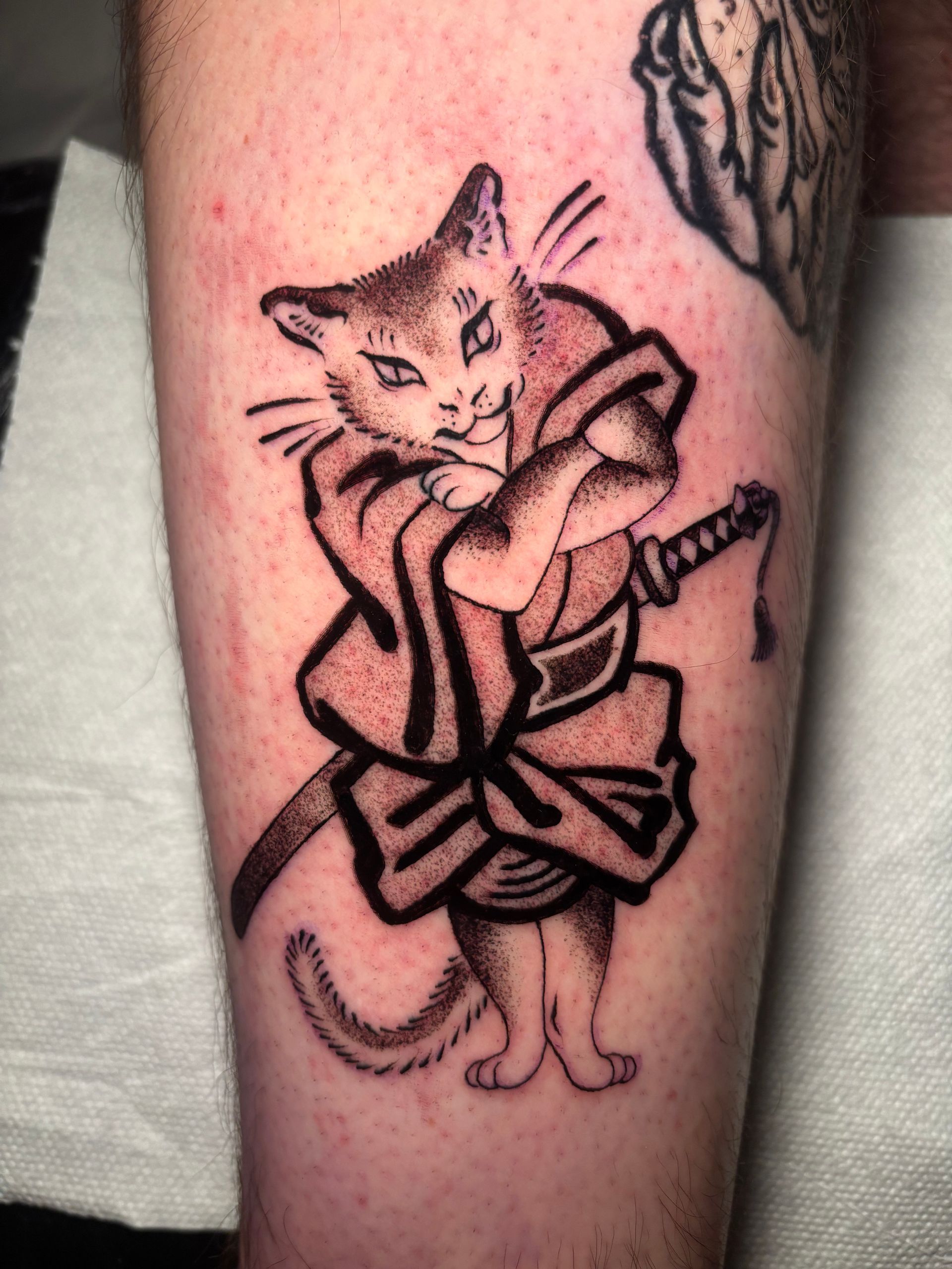 Tatuaje de un gato samurái con kimono y espada. Sombreado negro y gris en el brazo.