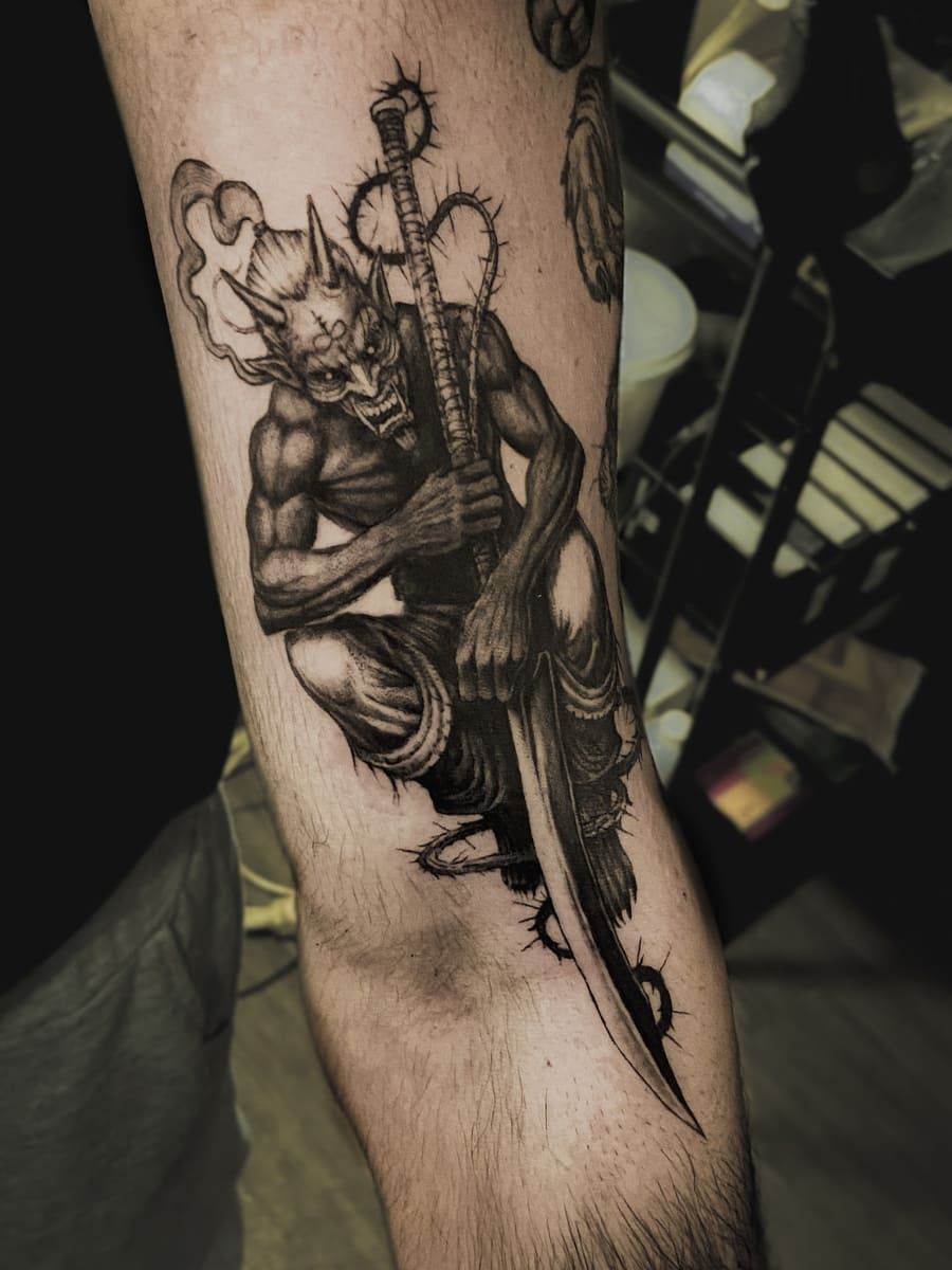 Tatuaje de un demonio con cuernos, sosteniendo una espada curva, en cuclillas sobre un brazo. Tinta en blanco y negro.