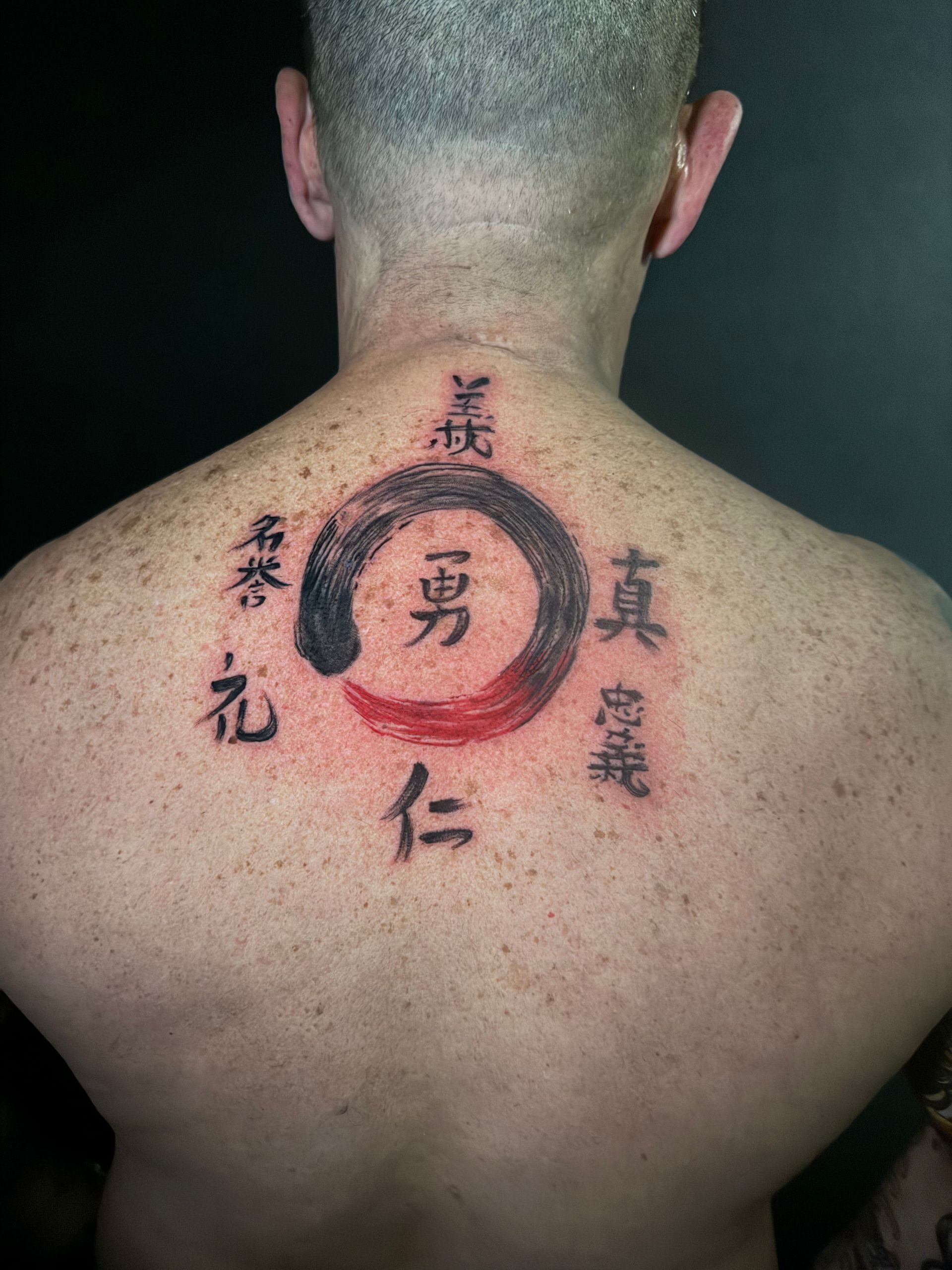 Tatuaje en la espalda: un círculo enso rojo y negro con caracteres kanji que representan el honor, el coraje y la verdad.