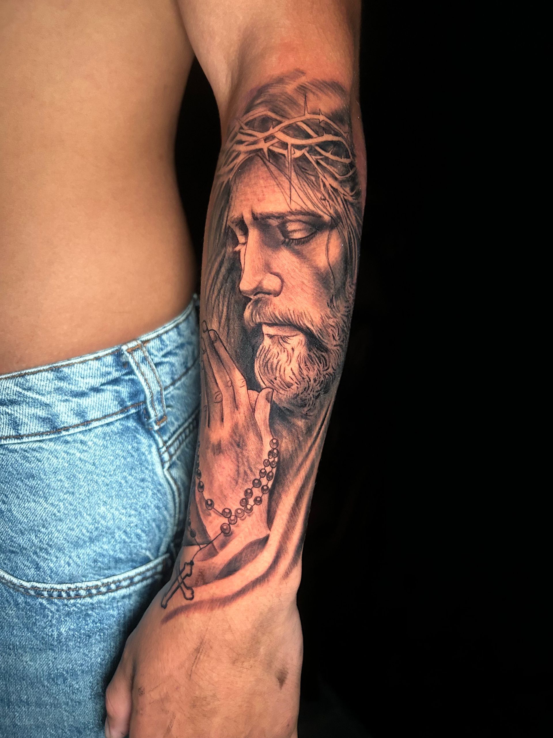 Tatuaje de Jesús en un antebrazo, portando una corona de espinas, mirando hacia abajo, con un rosario.