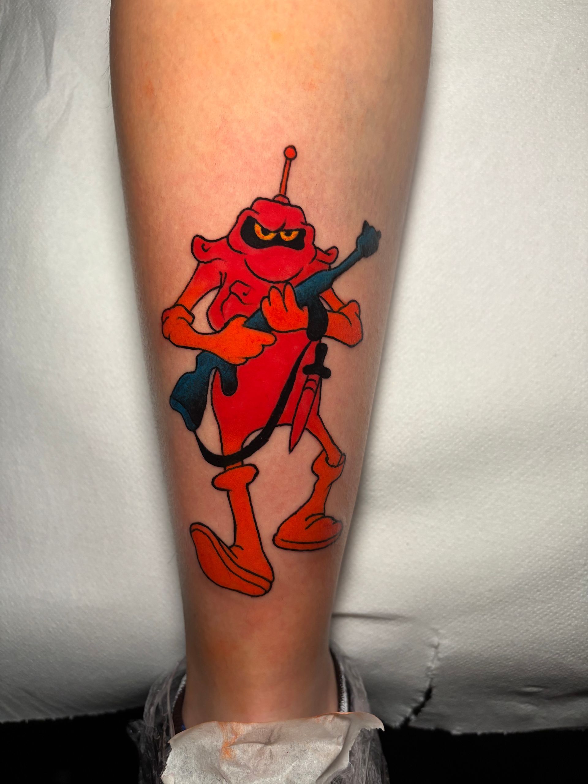 Tatuaje de robot de dibujos animados en el brazo, sosteniendo un rifle con expresión de enojo. Rojo y naranja con una pistola azul.