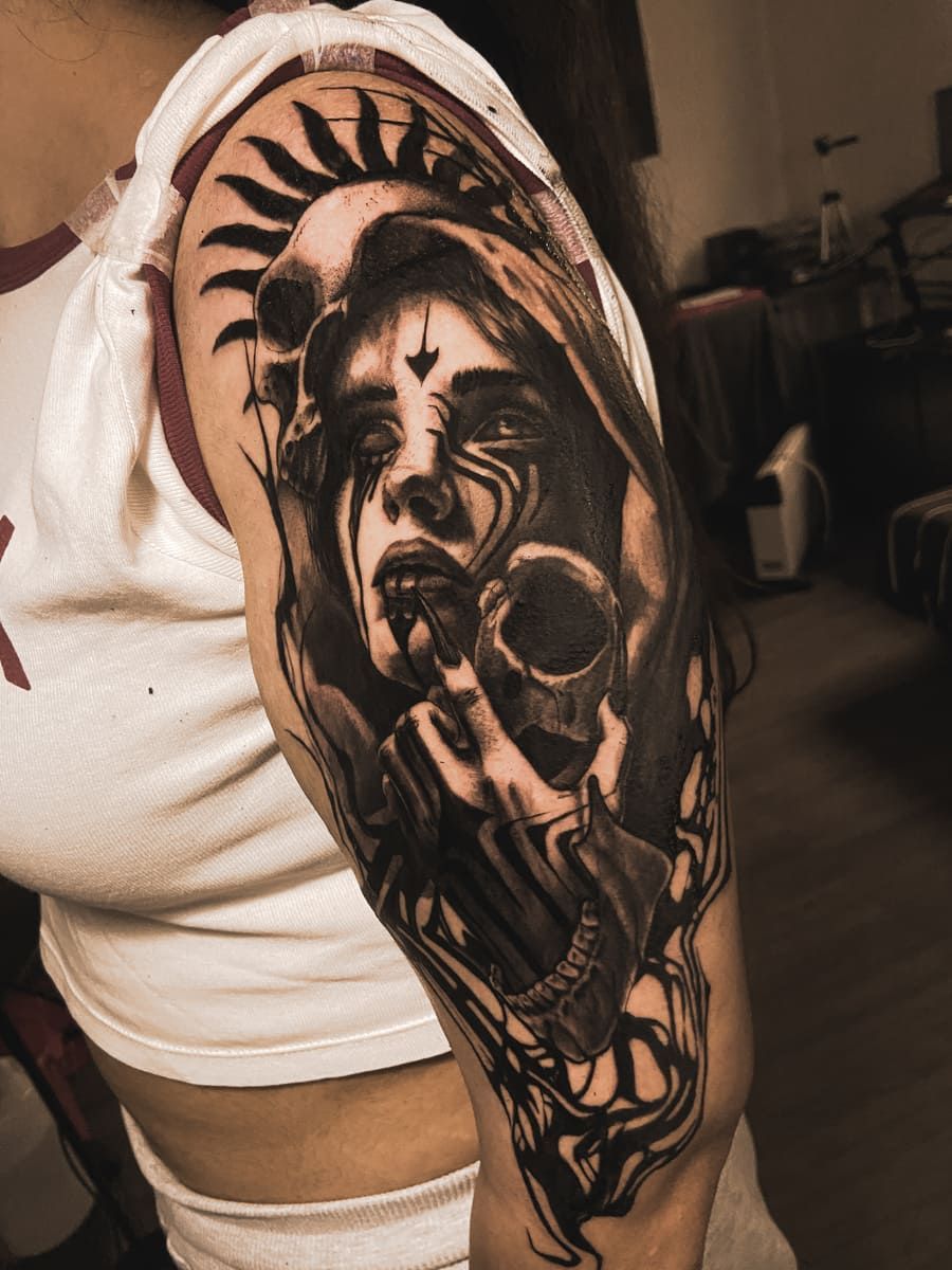 Tatuaje de una figura encapuchada sosteniendo una calavera. El dibujo es en blanco y negro y está en la parte superior del brazo. La figura parece estar llorando.