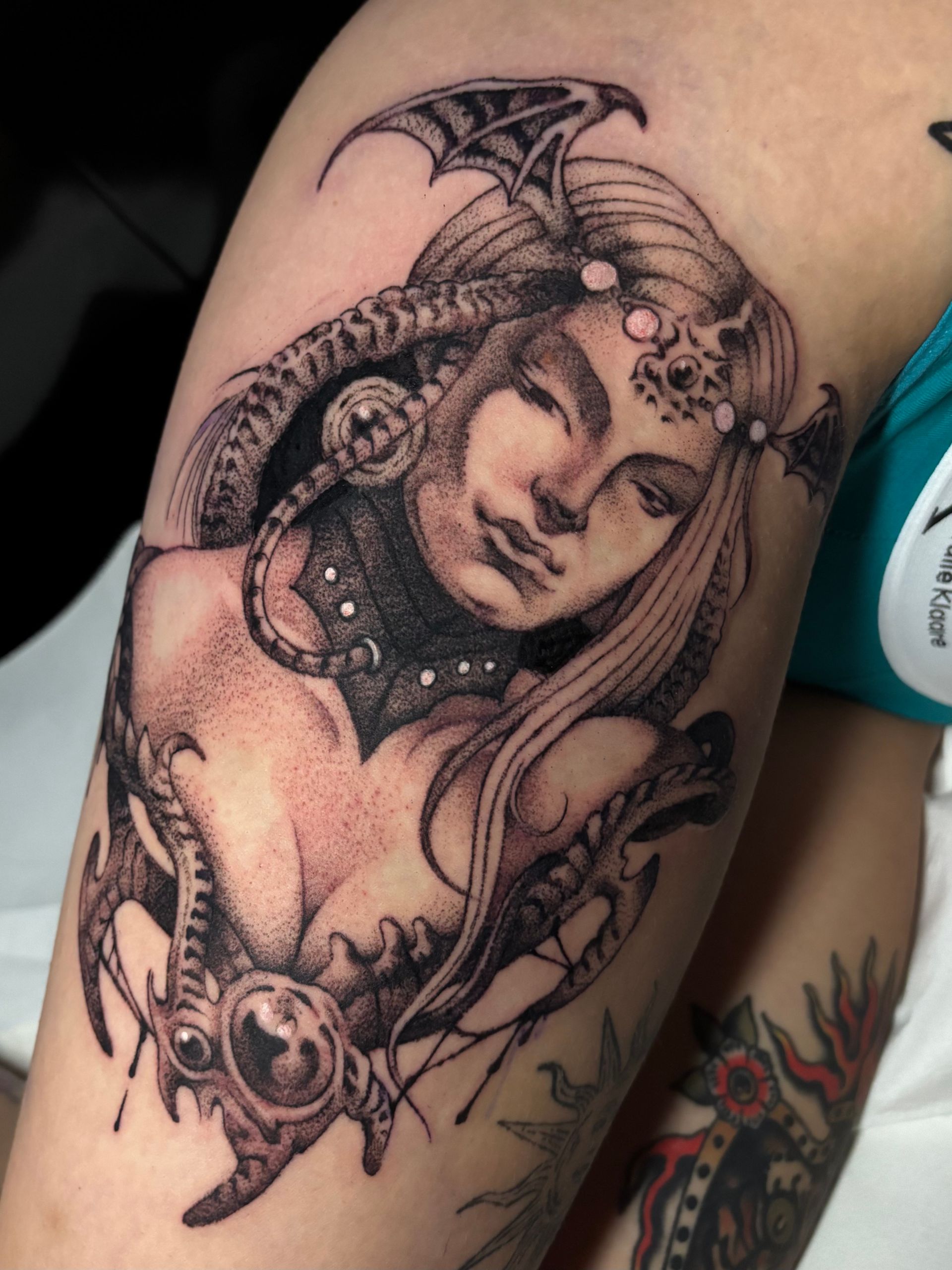 Tatuaje detallado en negro y gris de una mujer con rasgos de dragón, colocado en la parte superior del brazo de una persona.