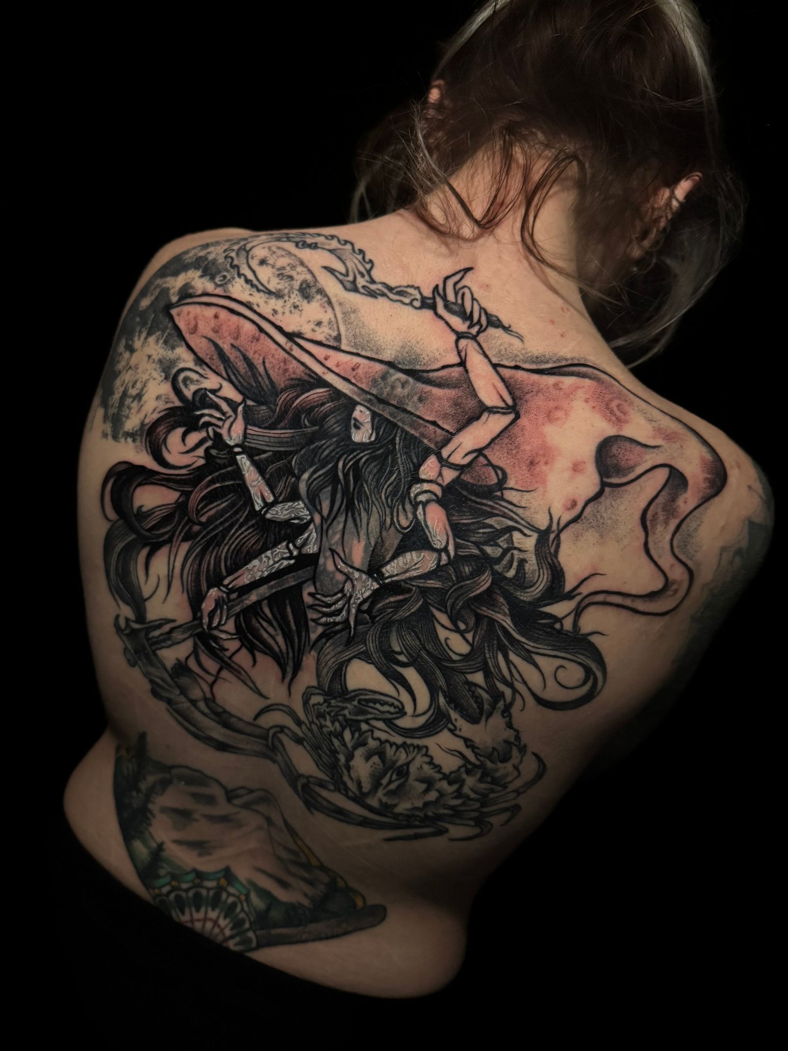 Espalda de mujer con un gran tatuaje de bruja negro y rojo, sosteniendo un bastón, rodeado de líneas en espiral.