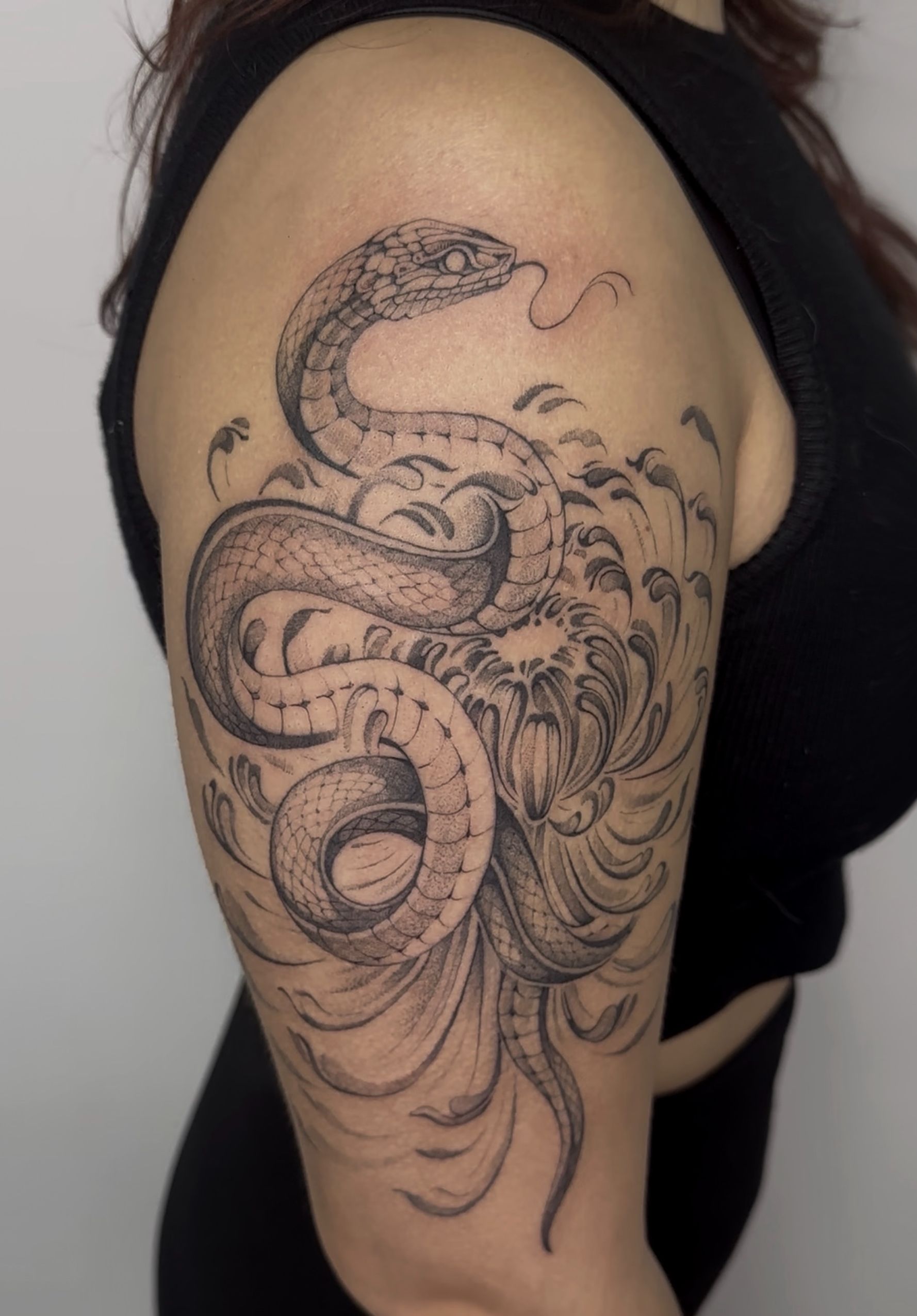 Tatuaje de una serpiente enrollada alrededor de una flor en la parte superior del brazo de una persona, en blanco y negro.