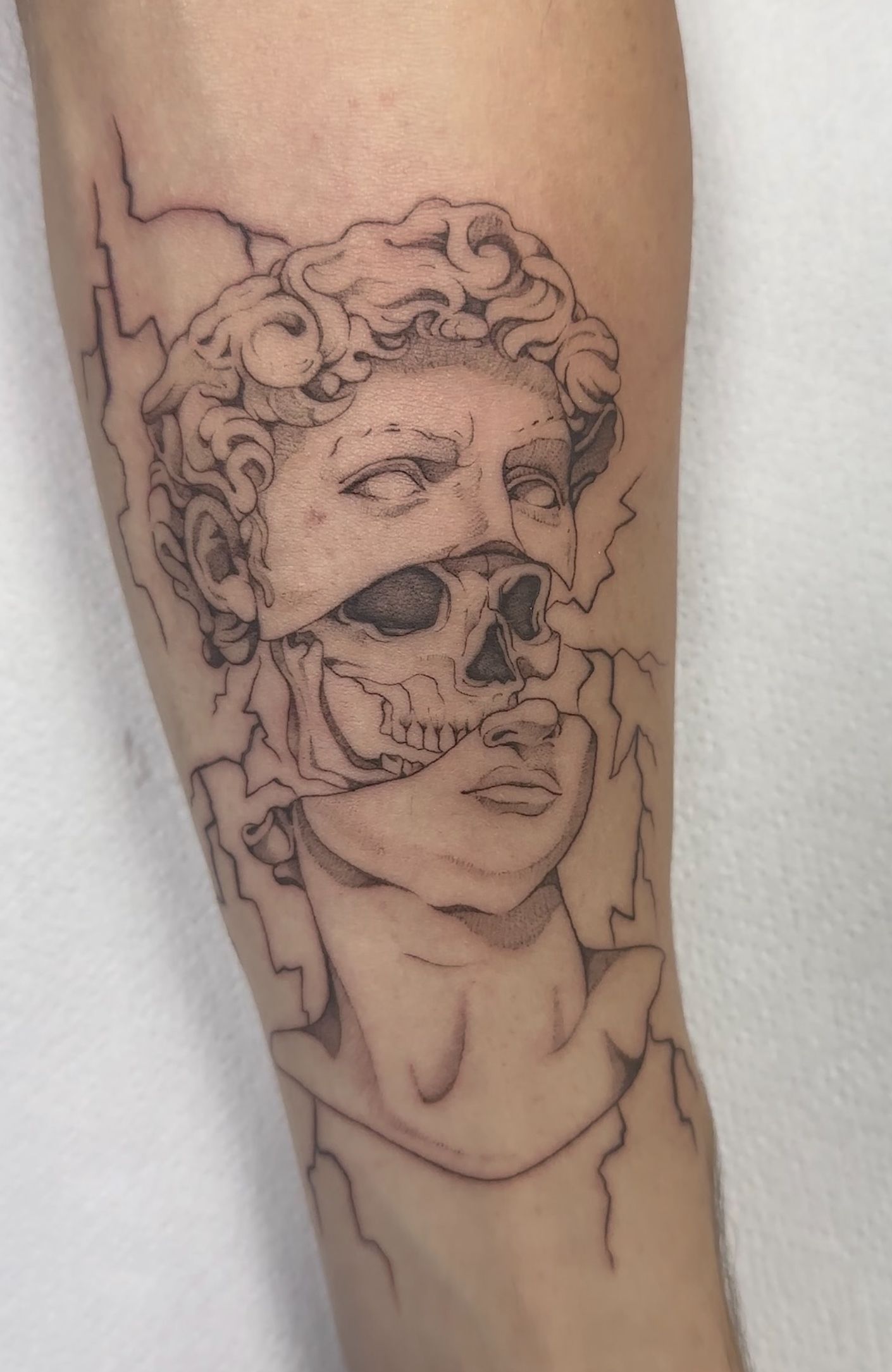 Tatuaje del busto de David con calavera al descubierto. Líneas grises con efecto agrietado en un brazo.