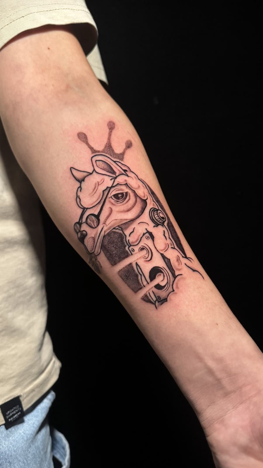 Tatuaje de una jirafa con una corona, con un contorno blanco y negro, en el antebrazo de una persona.