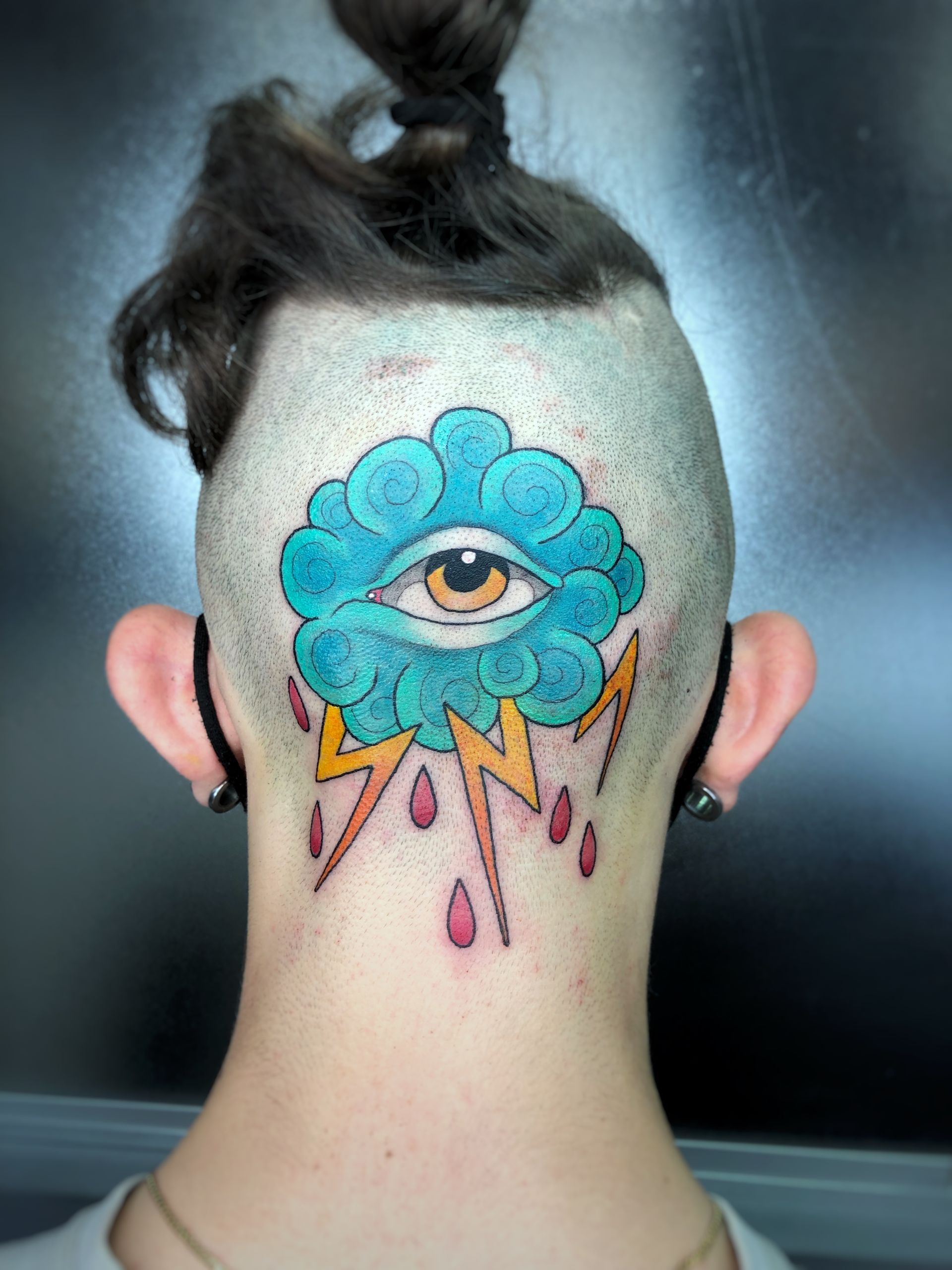 Tatuaje de una nube azul con un ojo, rayos y lluvia en la nuca rapada.