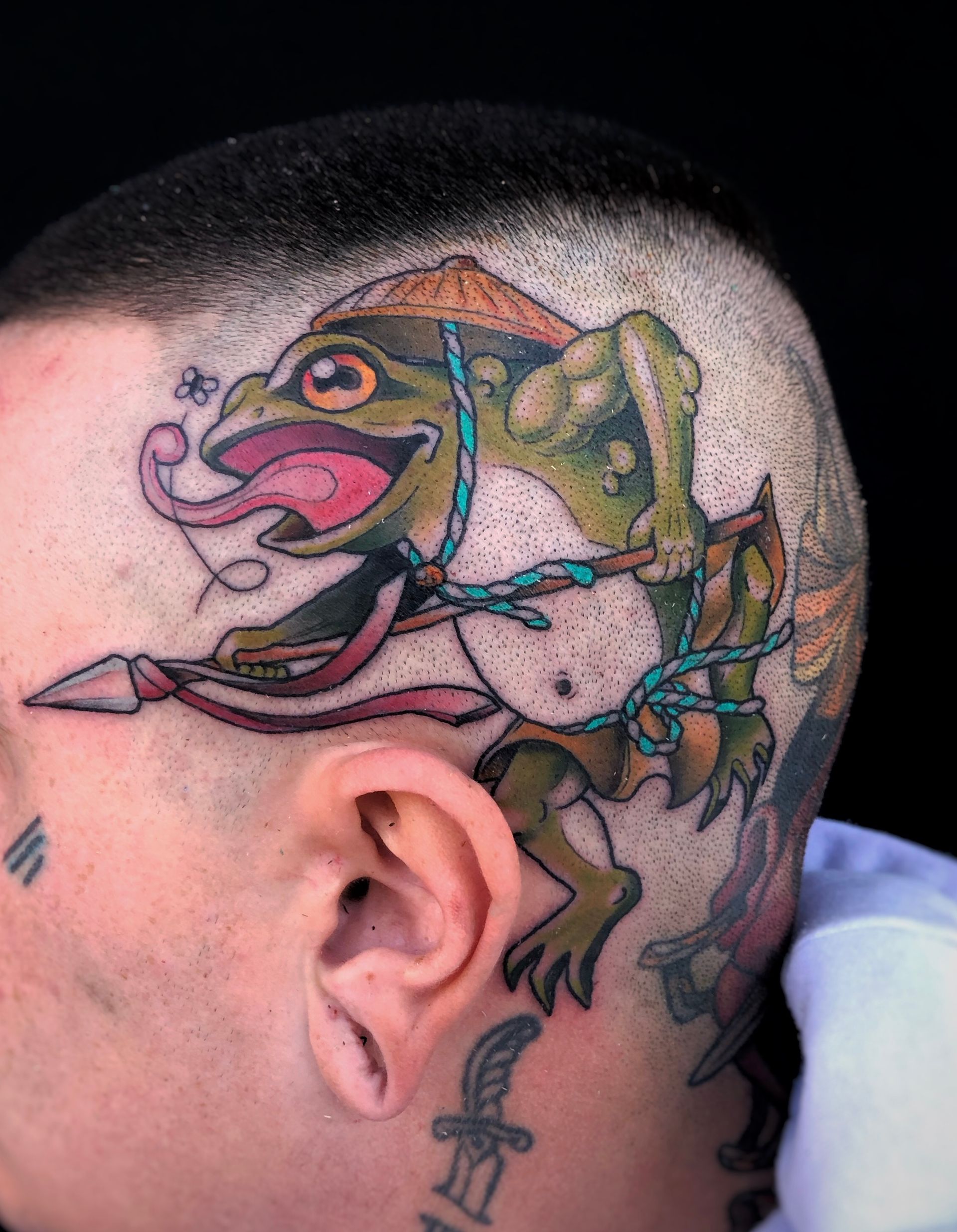 Tatuaje de una rana verde con un sombrero de paja sosteniendo una lanza, en la parte posterior de una cabeza rapada.