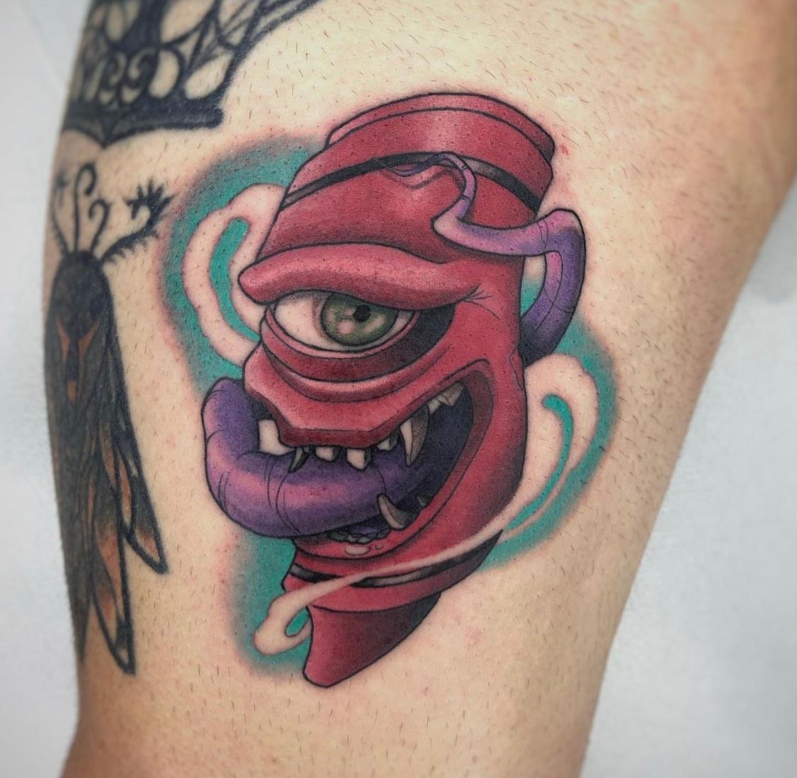 Tatuaje de un monstruo rojo, tuerto, con lengua y dientes morados en una pierna, rodeado de vapor azul y blanco.