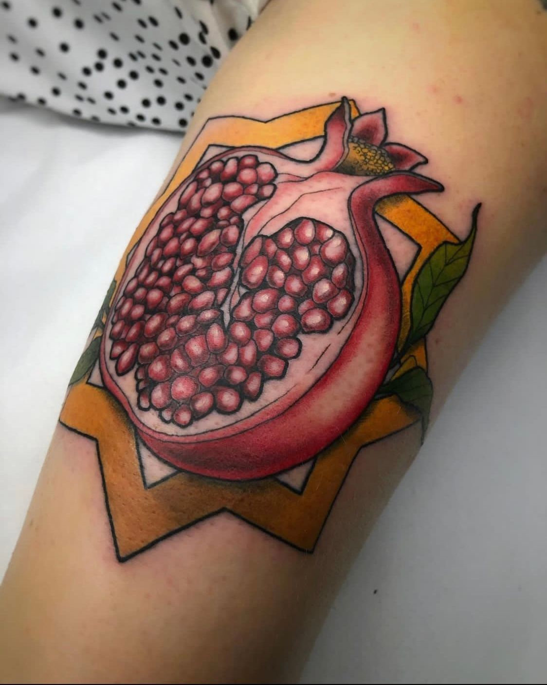 Tatuaje de una granada cortada, revelando semillas rojas, dentro de una forma de estrella amarilla sobre piel pálida.