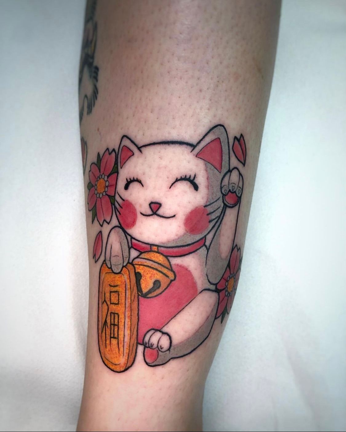 Tatuaje de un gato de la suerte Maneki-neko sonriente, rosa y gris, con una campana, flores y una pancarta.