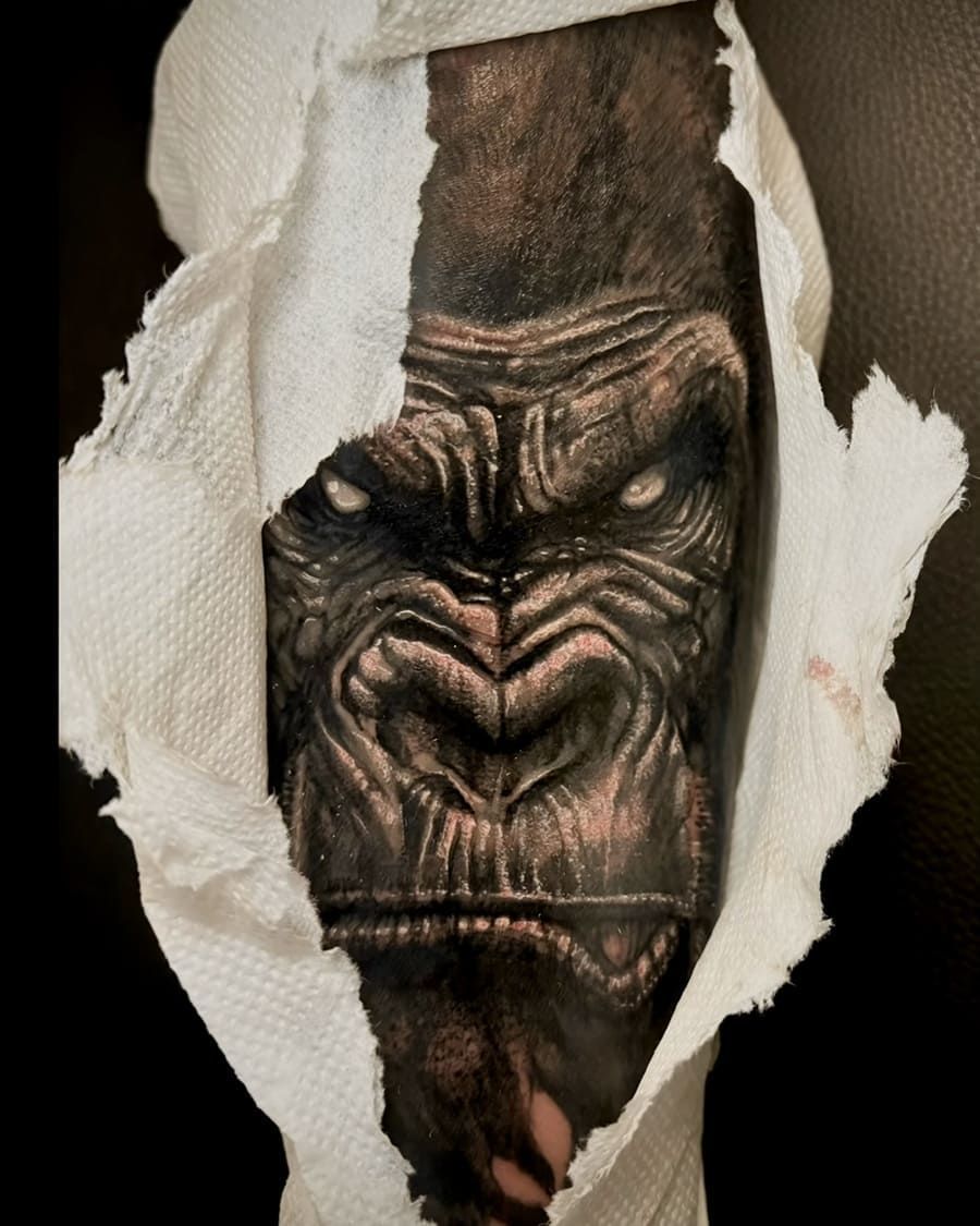 Tatuaje de cara de gorila que se asoma a través de papel de seda rasgado. Colores oscuros, mirada intensa.