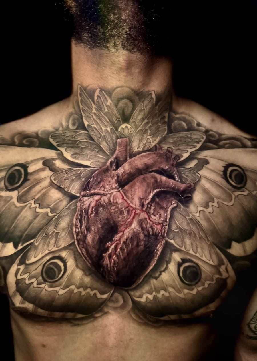 Tatuaje realista de un corazón en el pecho con alas de polilla. Cabello arcoíris. Fondo oscuro.