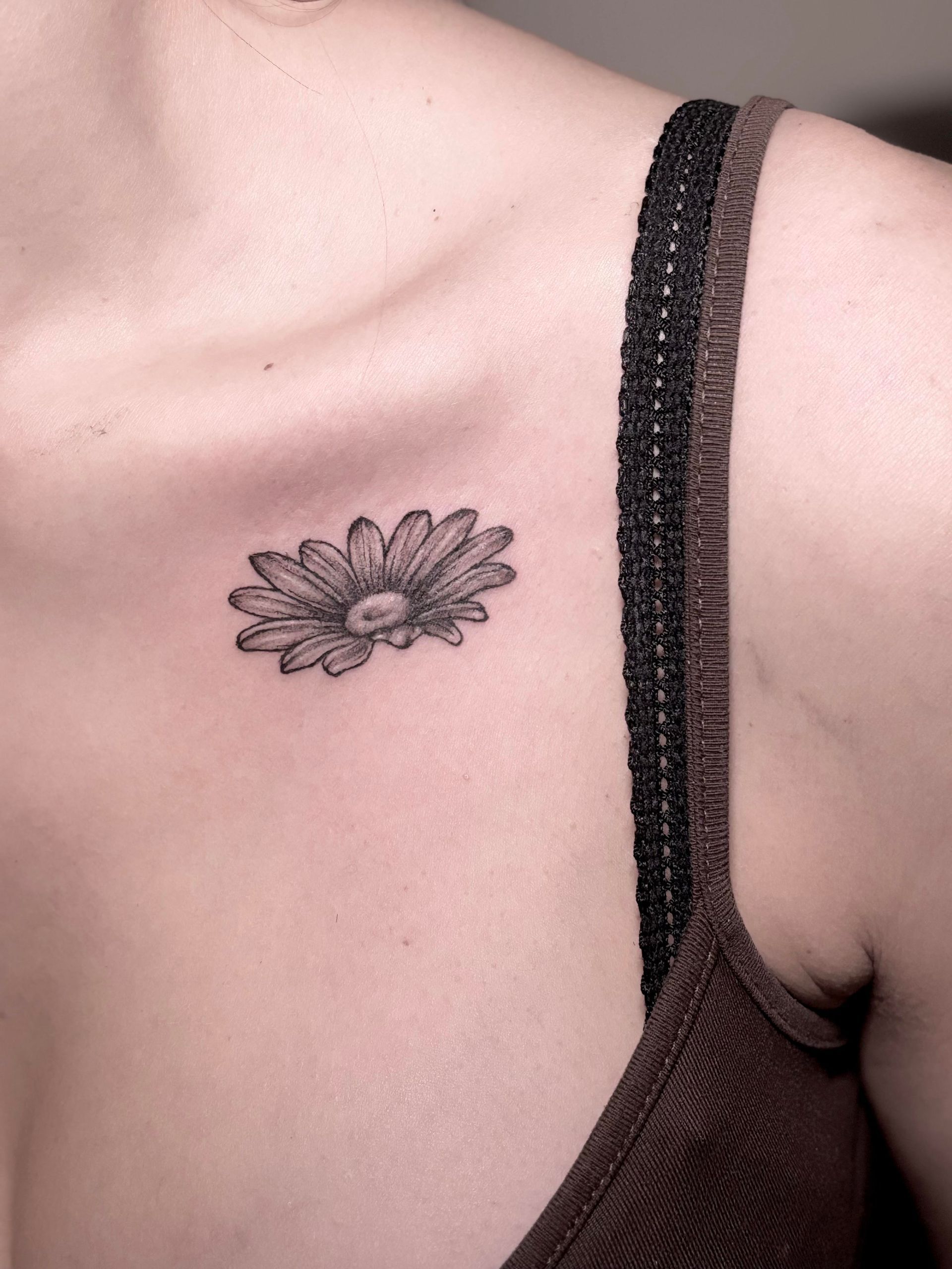 Tatuaje de una margarita en el pecho de una persona, al lado de una tira de una camiseta marrón.