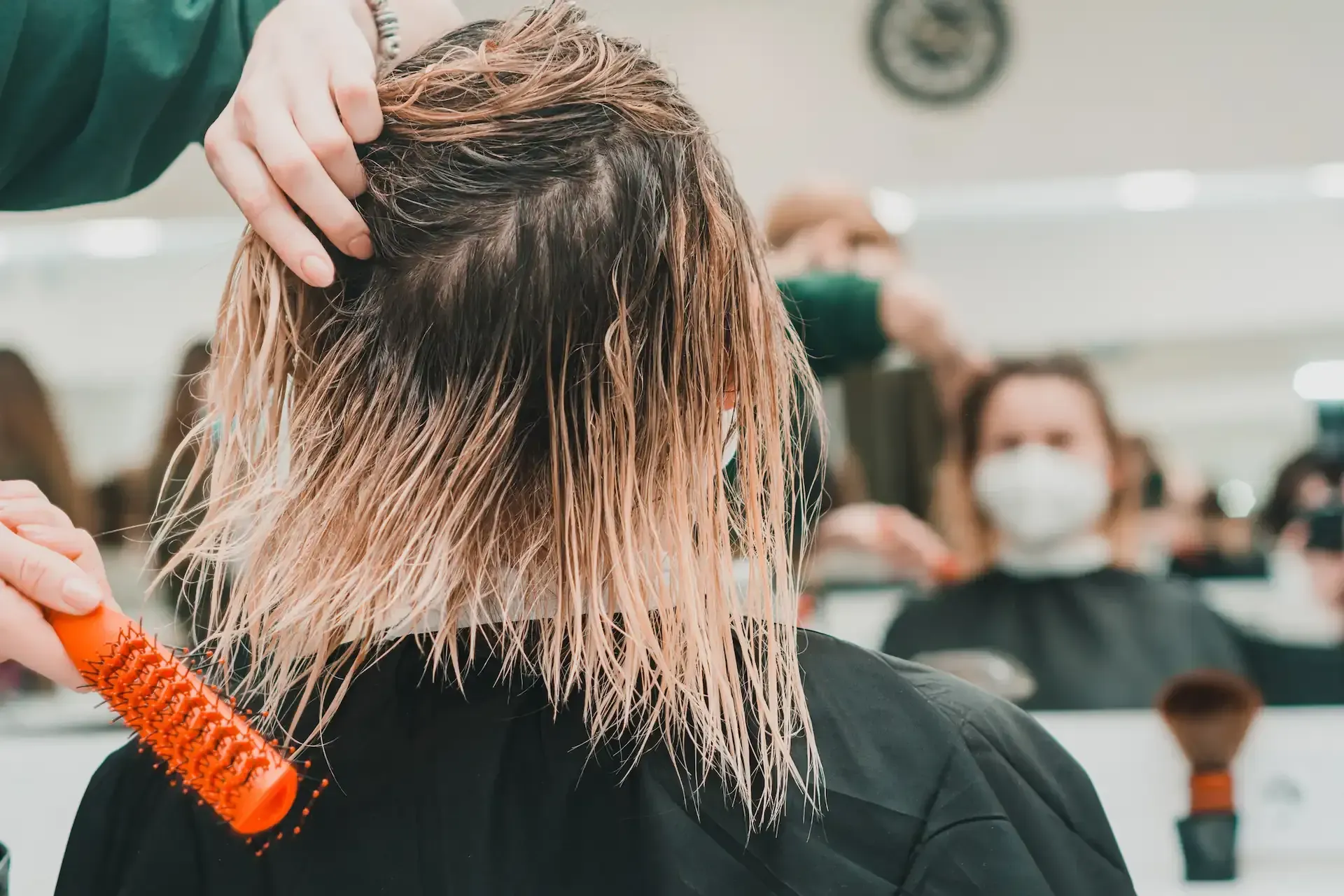 Una peluquera peina el cabello mojado en un salón. Hay otras personas al fondo.