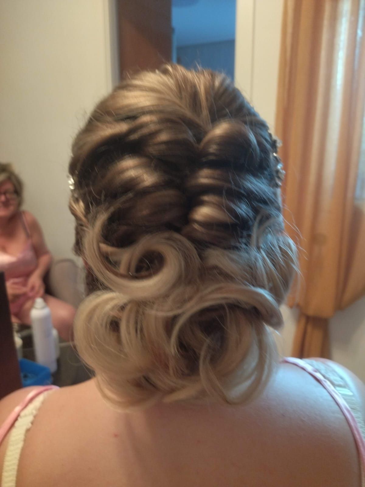 Cabello de mujer peinado en un recogido con rizos rubios y secciones trenzadas más oscuras; hay alguien en el fondo.