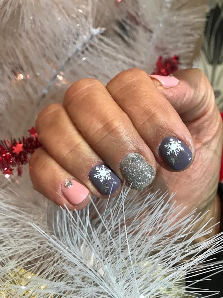 Mano con uñas pintadas en tonos rosa, gris y brillo, con diseños de copos de nieve, sobre un fondo de árbol blanco.