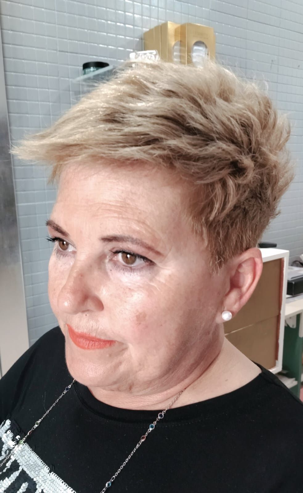 Mujer con cabello rubio corto y puntiagudo, vestida con una camisa negra y aretes de perlas, mirando hacia un lado.