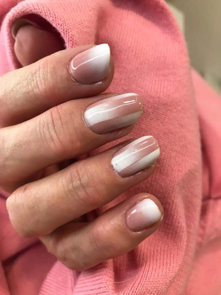 Mano con uñas cortas pintadas con diseño ombré y rayas en tonos blanco y nude.