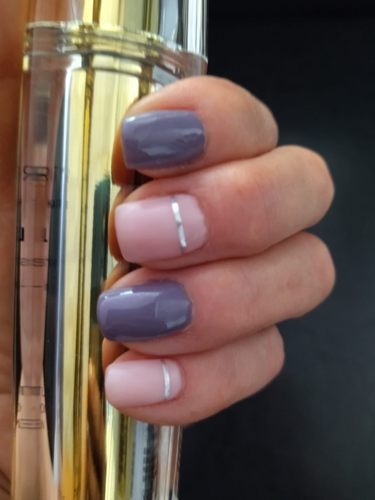 Mano con esmalte de uñas morado y rosa, una uña tiene una raya plateada, sosteniendo un objeto dorado.