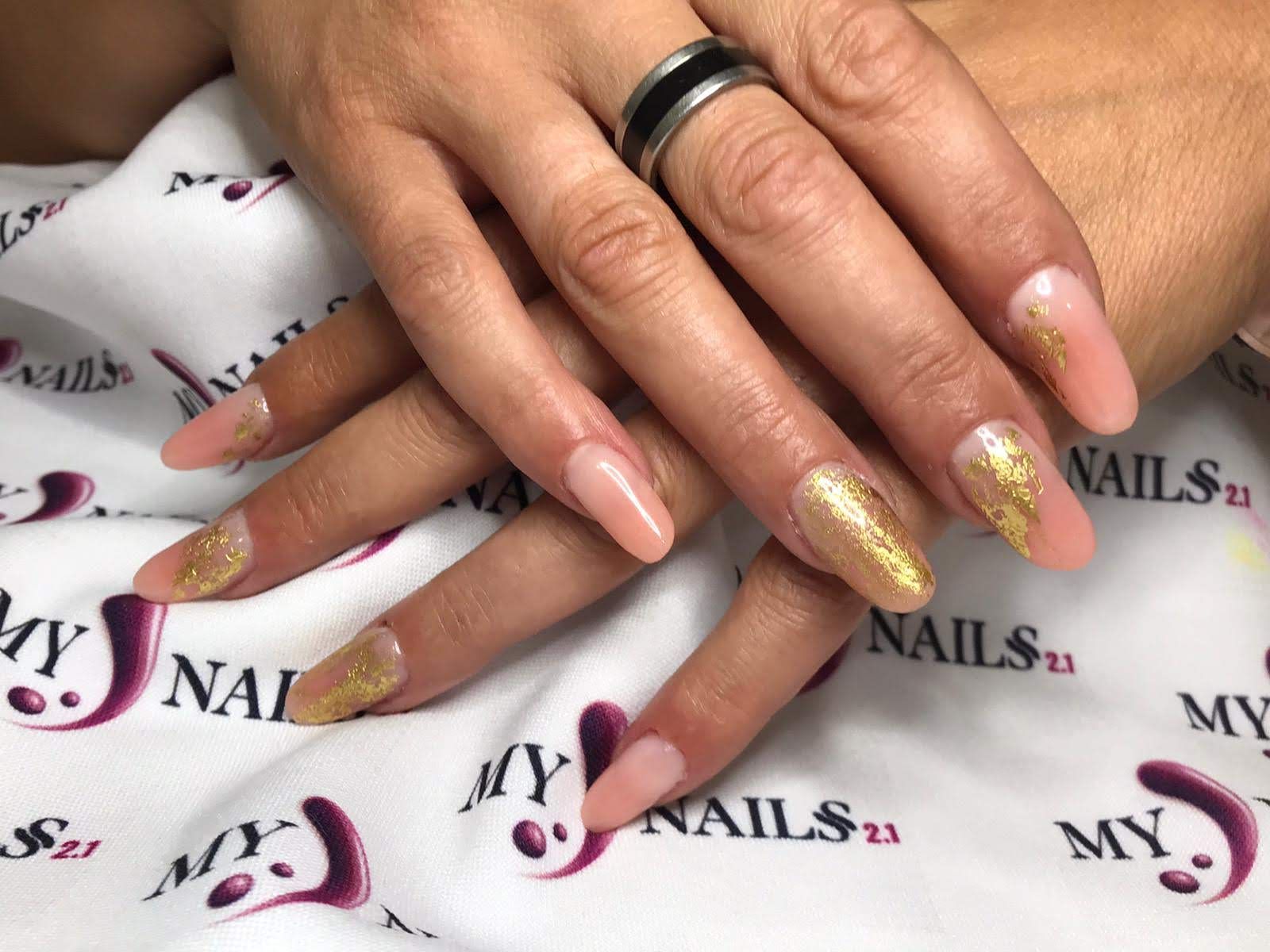 Manos con brillo dorado y uñas acrílicas color nude, anillo en un dedo, apoyado sobre una superficie blanca con logo impreso.