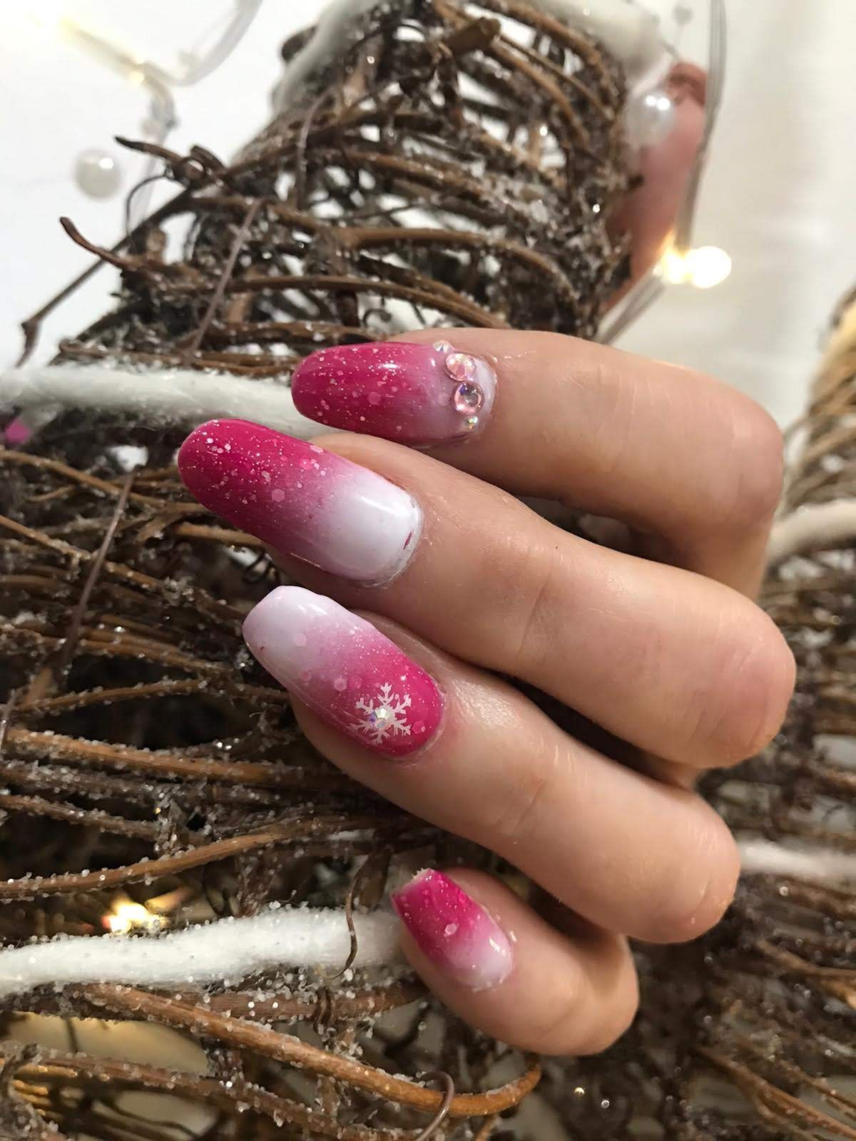 Arte de uñas en degradado rosa y blanco con detalles de copos de nieve y joyas, mano apoyada sobre una corona tejida.