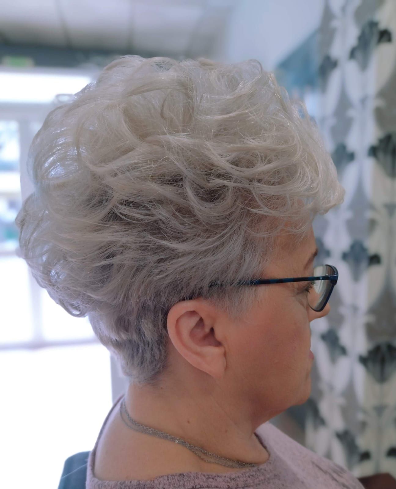Mujer con cabello corto, en capas, gris y rizado, con gafas, mirando hacia un lado.