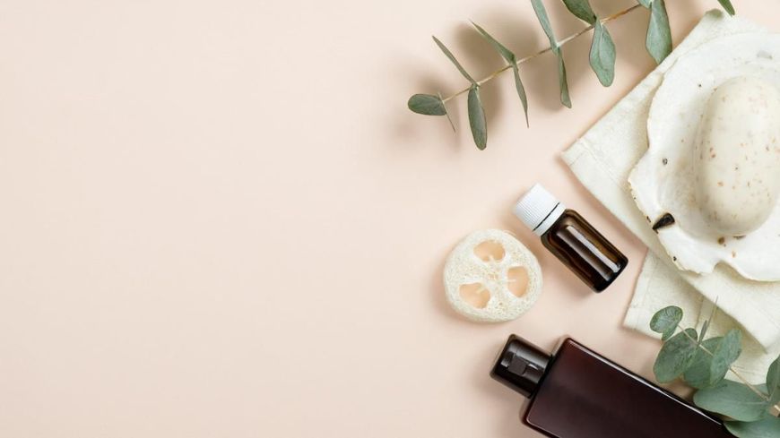 Una toma minimalista desde arriba de artículos esenciales para un spa: una botella marrón, aceite esencial, lufa y jabón sobre una concha con eucalipto.