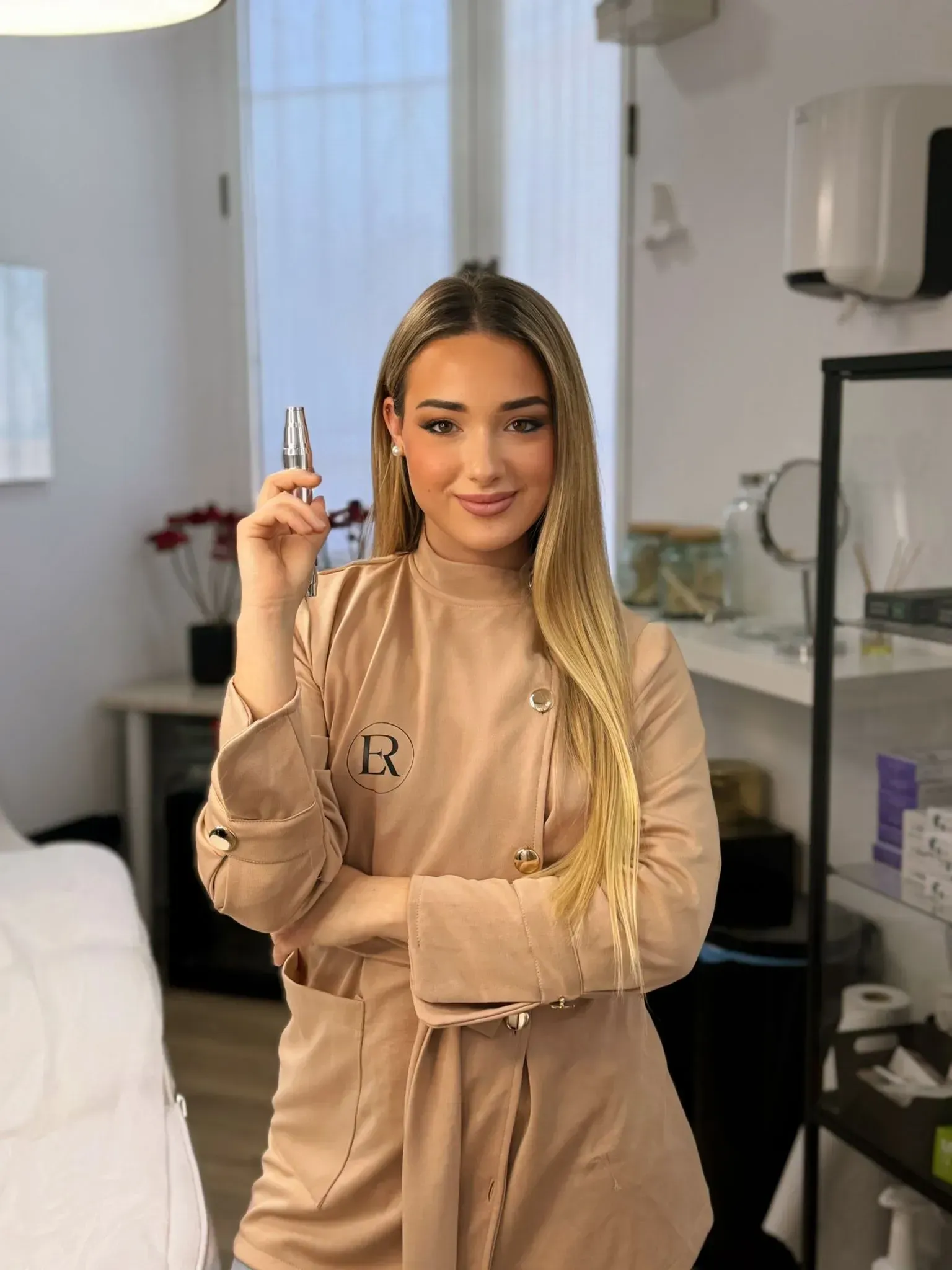 En una clínica luminosa, una persona  viste un uniforme beige y sonríe mientras sostiene un pequeño frasco de cosméticos.