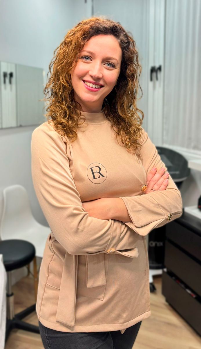 Una persona con cabello rizado sonríe mientras permanece de pie con los brazos cruzados en un espacio de oficina luminoso y moderno.