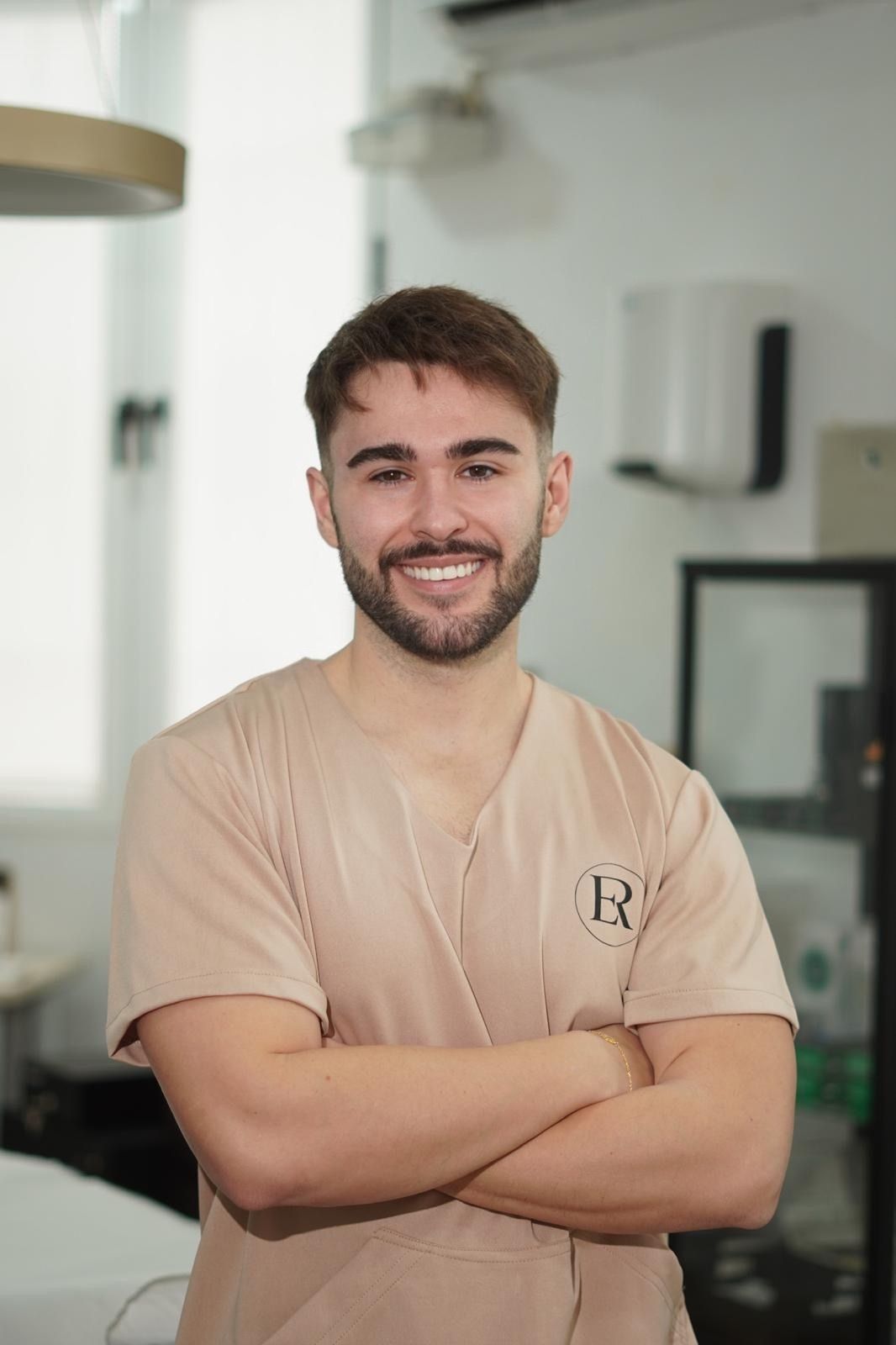 Una persona con barba y cabello castaño corto, vestida con una bata médica de color beige y con los brazos cruzados