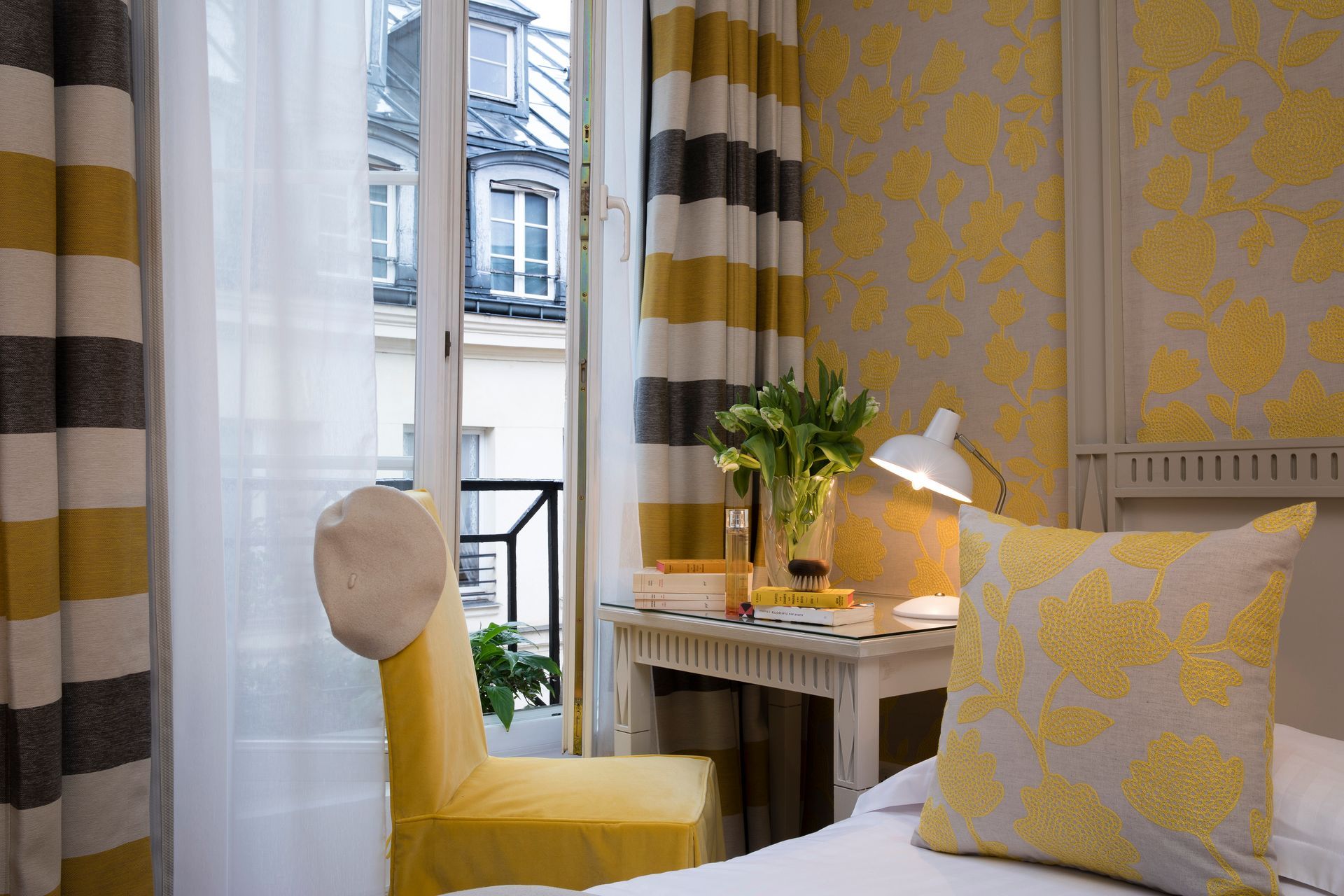 Chambre avec des touches de jaune : rideaux à rayures, papier peint fleuri, bureau, vue depuis le balcon et chaise jaune.