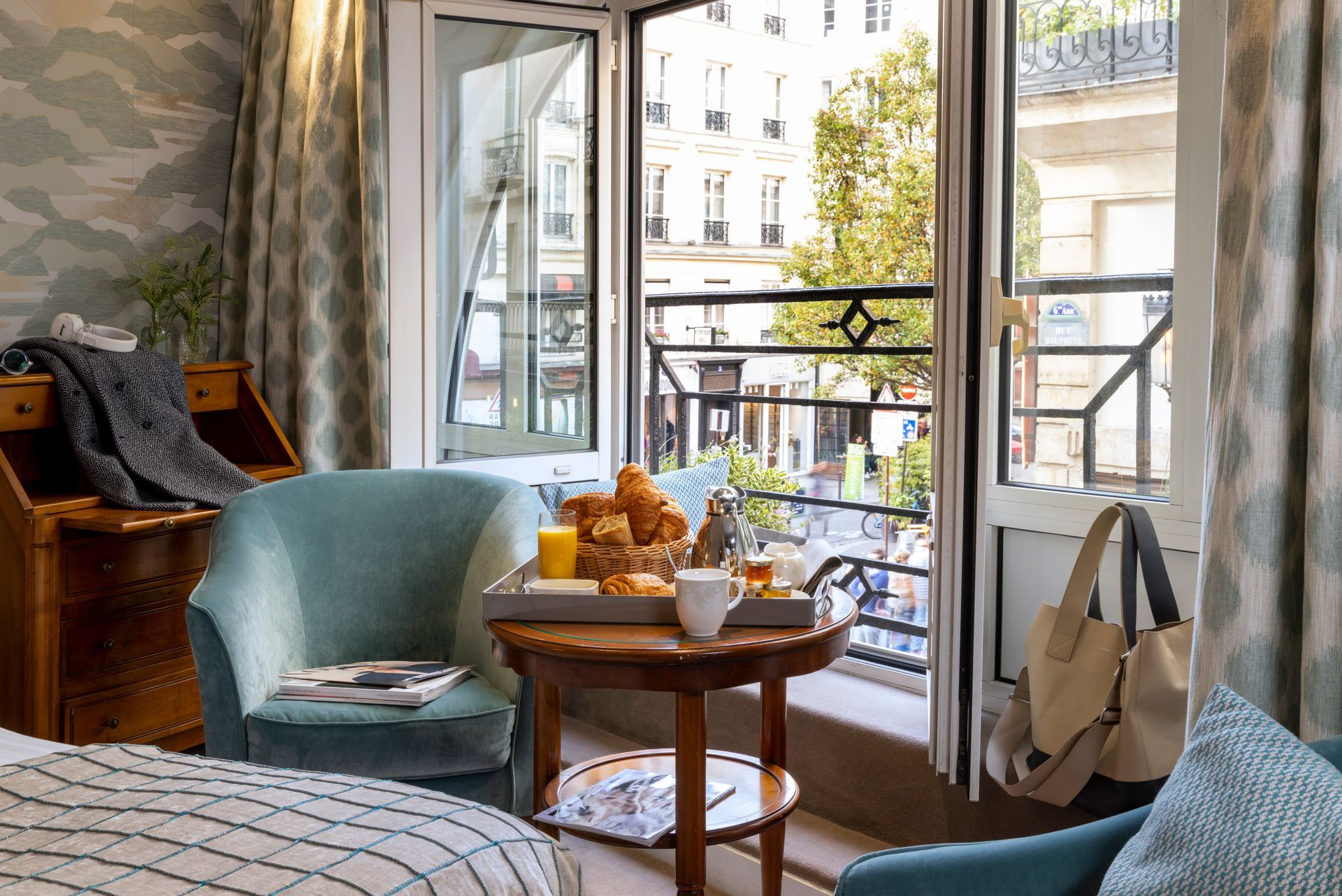 Chambre d'hôtel avec portes-fenêtres ouvertes sur le balcon, table dressée pour le petit-déjeuner avec croissants, donnant sur une rue de la ville.