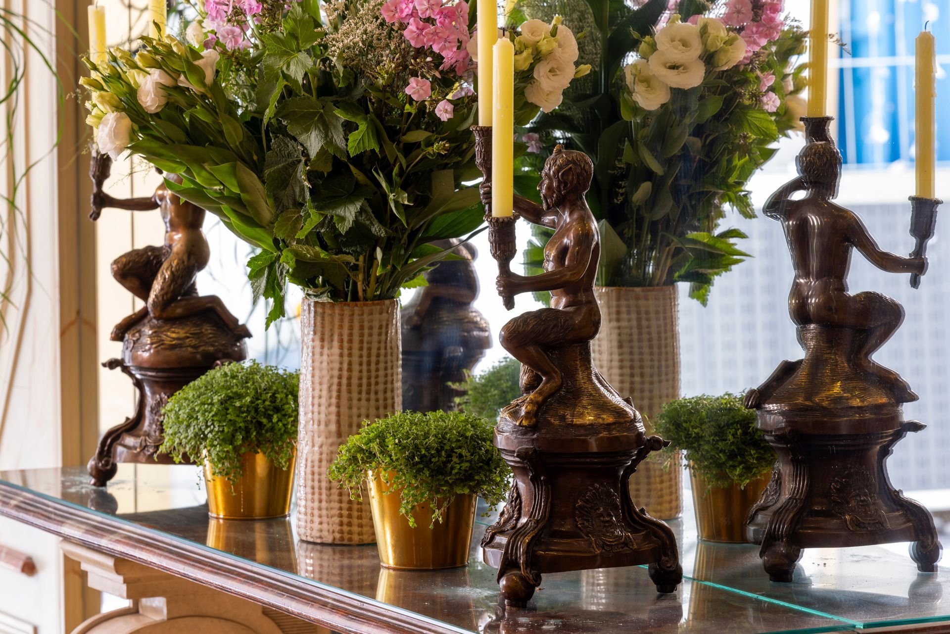 Des bougeoirs en bronze en forme de sirène avec des fleurs sur une table, des pots dorés remplis de verdure, des miroirs.